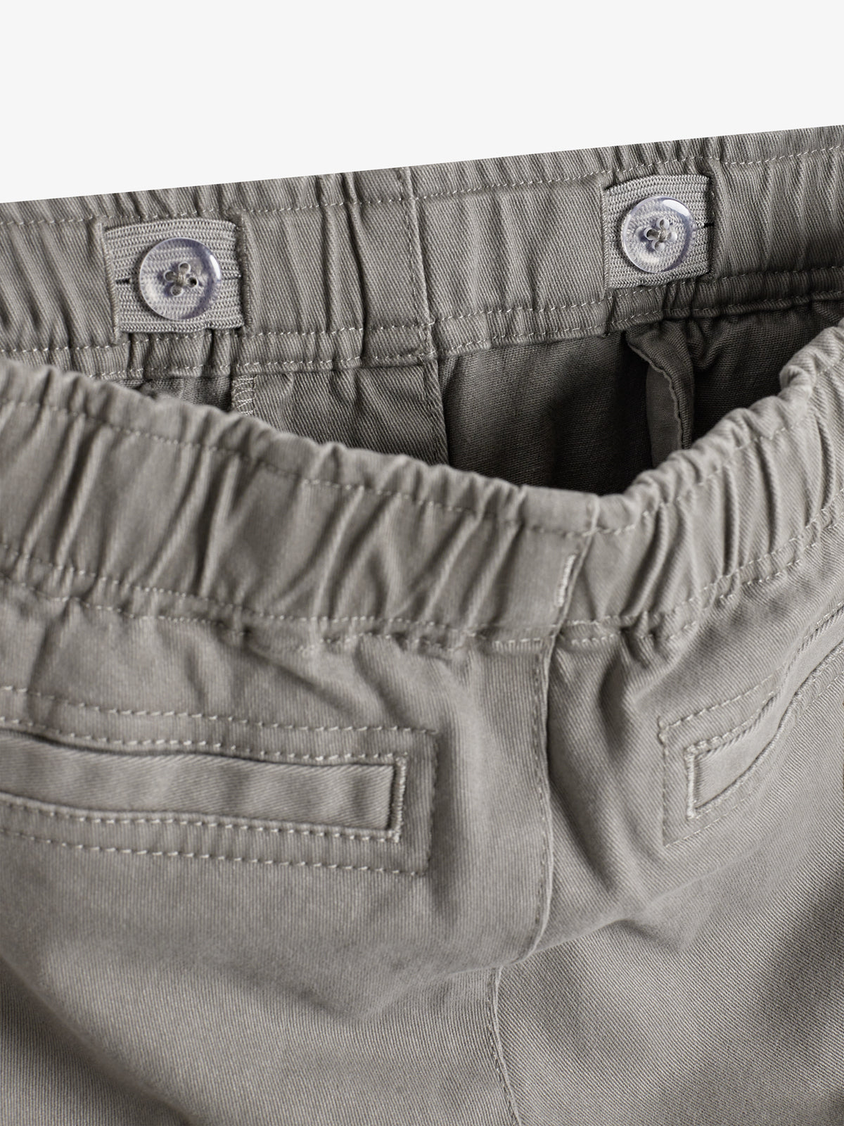 Grijze broek met elastische tailleband en knopen, hangt ontspannen. Twee steekzakken aan de achterkant zichtbaar. In een eenvoudige, goed verlichte omgeving tegen een witte achtergrond.
