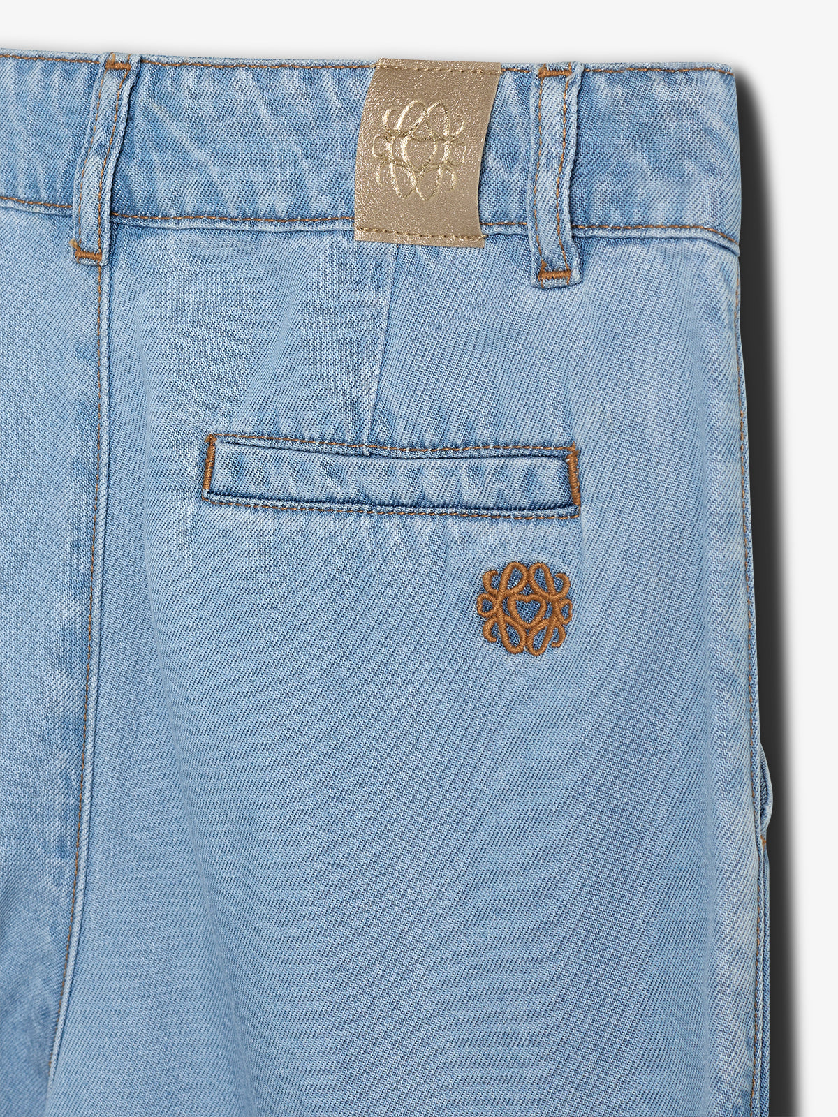 Een lichtblauwe spijkerbroek met een geborduurd, bruin logo op de zak en een beige leren label op de tailleband. Lichtbruin stiksel accentueert de zakken en riemlussen.