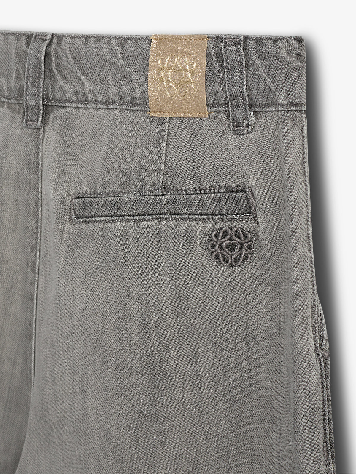 Grijze broek met een gespierde tailleband en een achterzak in een lichte omgeving. Een lichtbruine leren patch toont een geborduurd embleem. Geen tekst aanwezig.