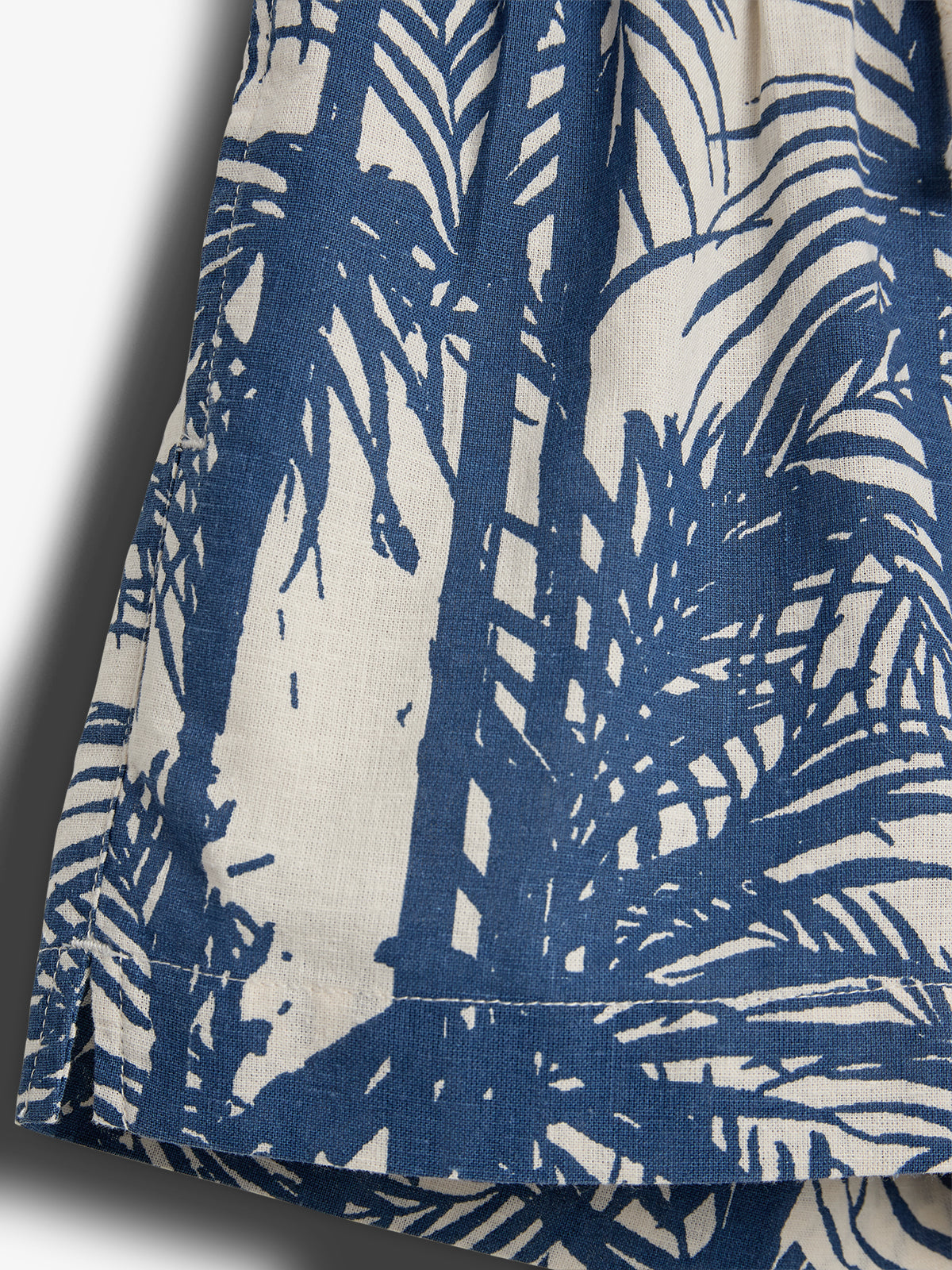 Stof met blauw-witte palmboomprint. Geen tekst aanwezig. De stof hangt los, met licht en schaduweffecten aan de linkerkant.
