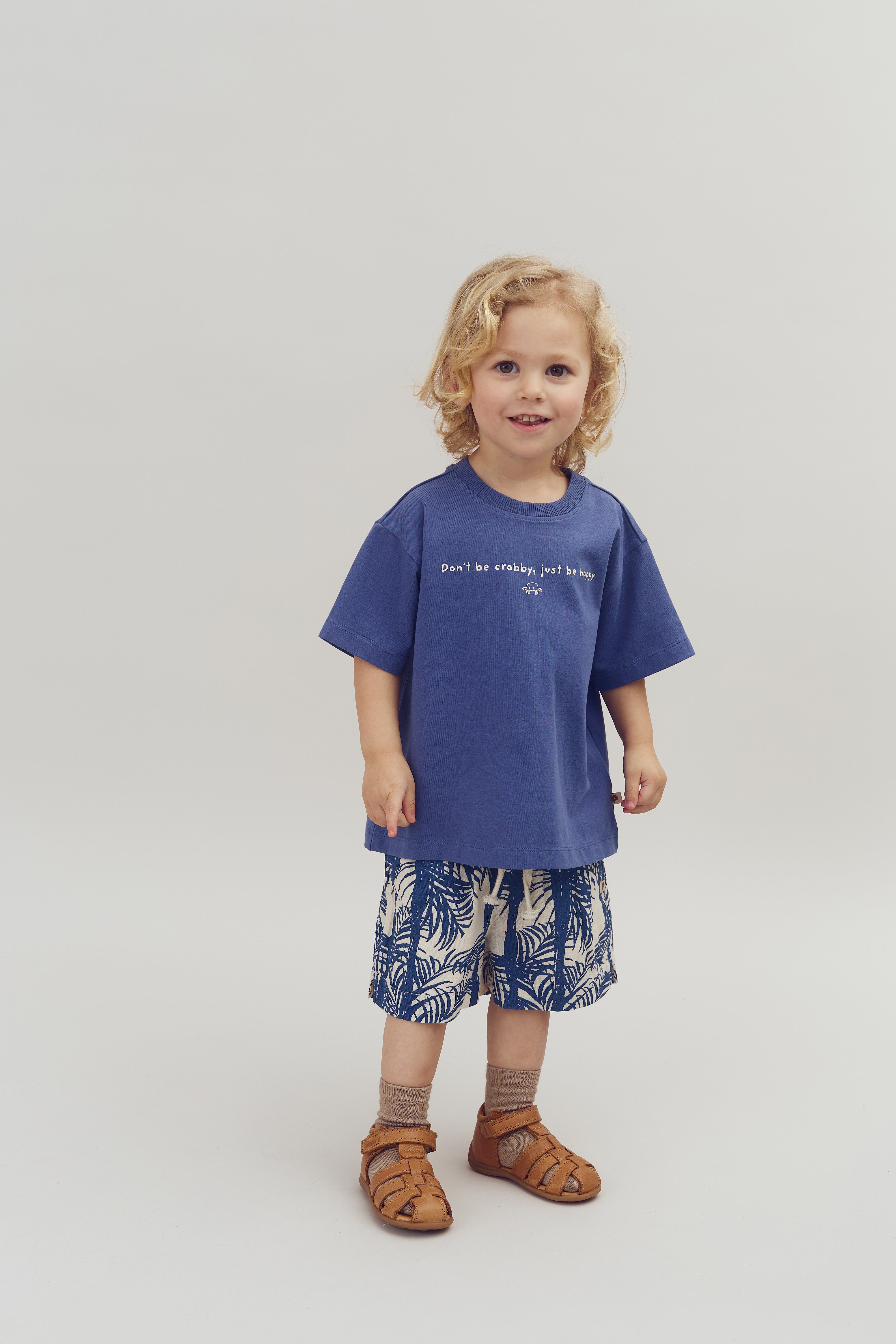 Een kind draagt een blauw T-shirt met de tekst "Don't be crabby, just be happy" en een kleine krab, gecombineerd met korte broek en sandalen, in een neutrale studio-omgeving.