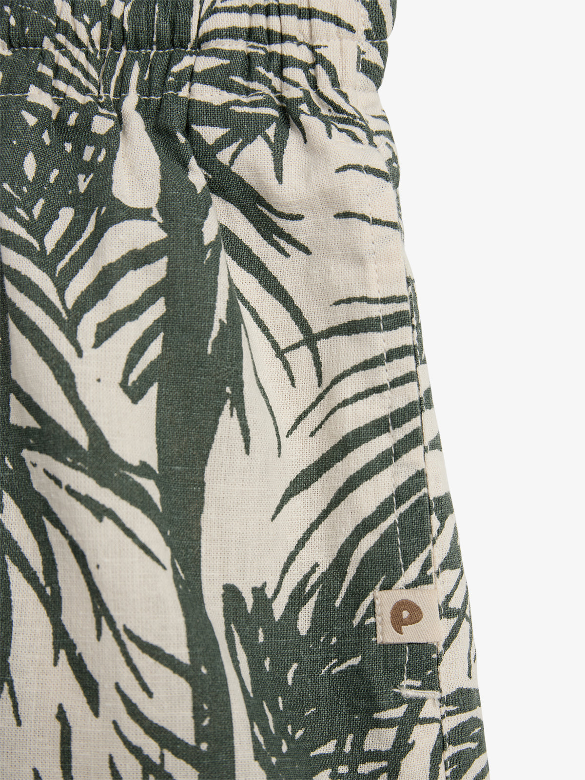 Een kledingstuk met een print van groene bladeren op een beige achtergrond. Een elastische tailleband is zichtbaar. Een klein label met een "P" bevindt zich aan de zijkant.