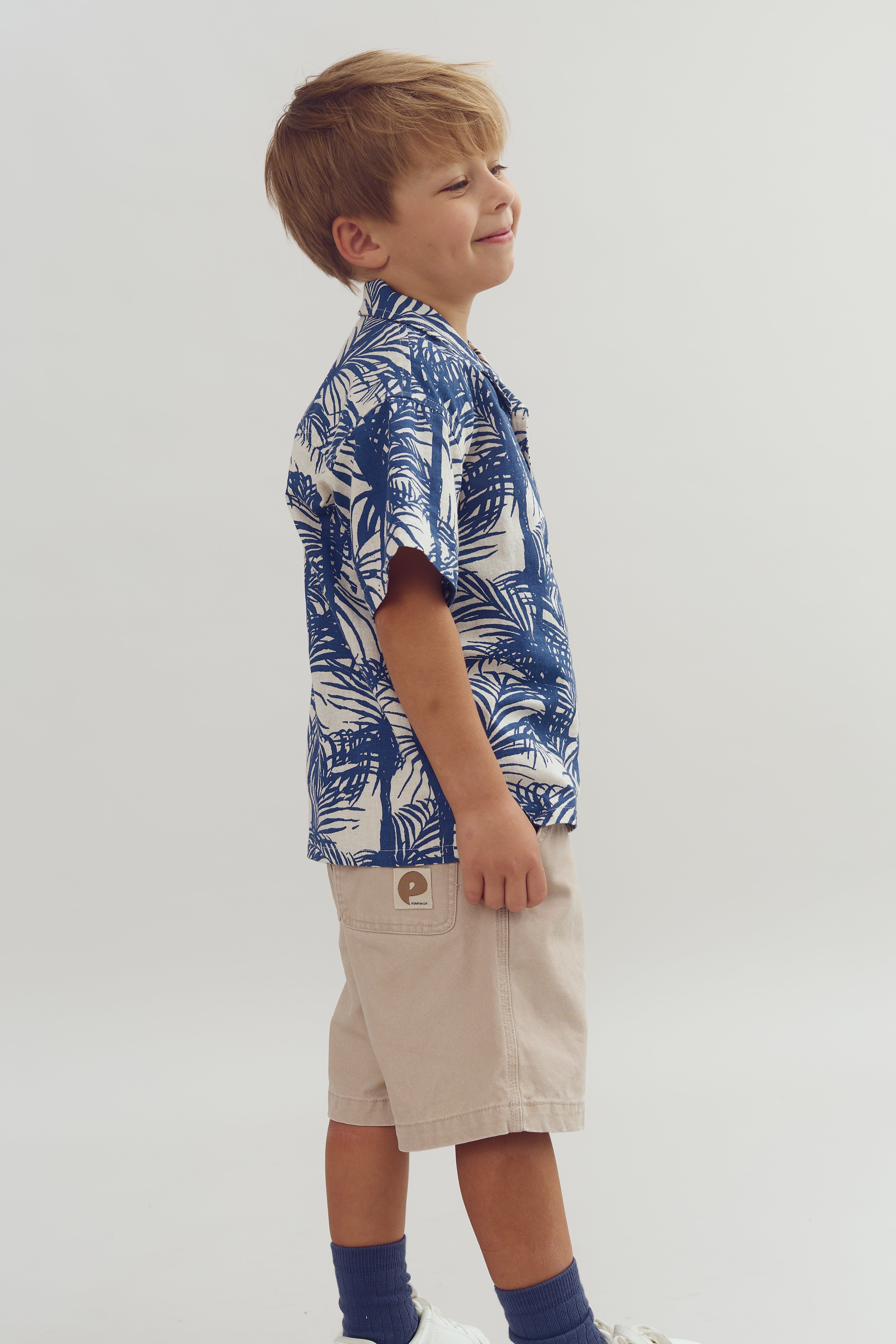 Een jong kind draagt een blauw-wit tropisch shirt en beige korte broek, glimlachend in een neutrale, studio-omgeving. Er is een embleem met een "e" op de broekzak.