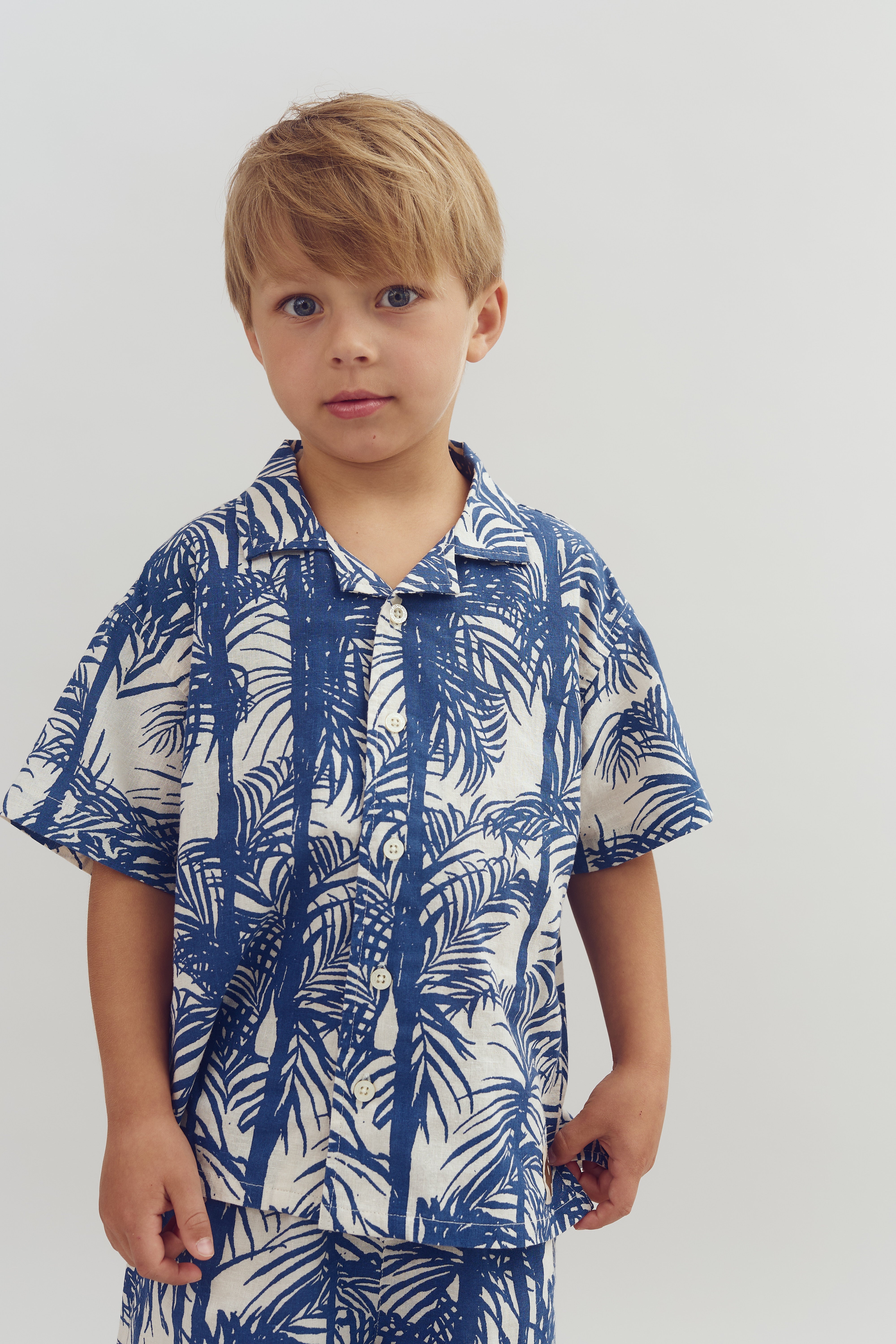 Een jongen draagt een overhemd met blauwe palmbomenprint, staand tegen een effen, witte achtergrond. Zijn blik is neutraal en ontspannen.