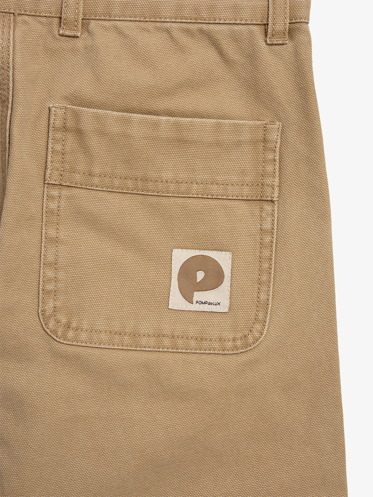 A khaki trouser pocket features a sewn label with a brown logo resembling a "P" and the text "POMPdeLUX" on a simple, plain background.