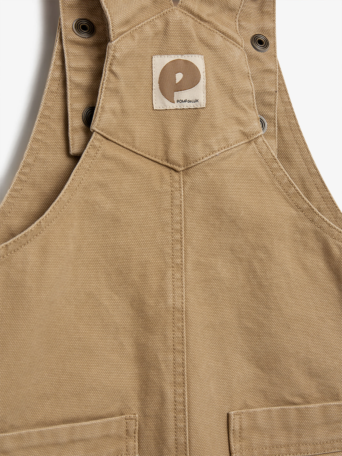 A beige fabric bib with metal buttonholes, featuring a small patch displaying a stylized "P" and the text "POMPDEUX," set against a plain background.