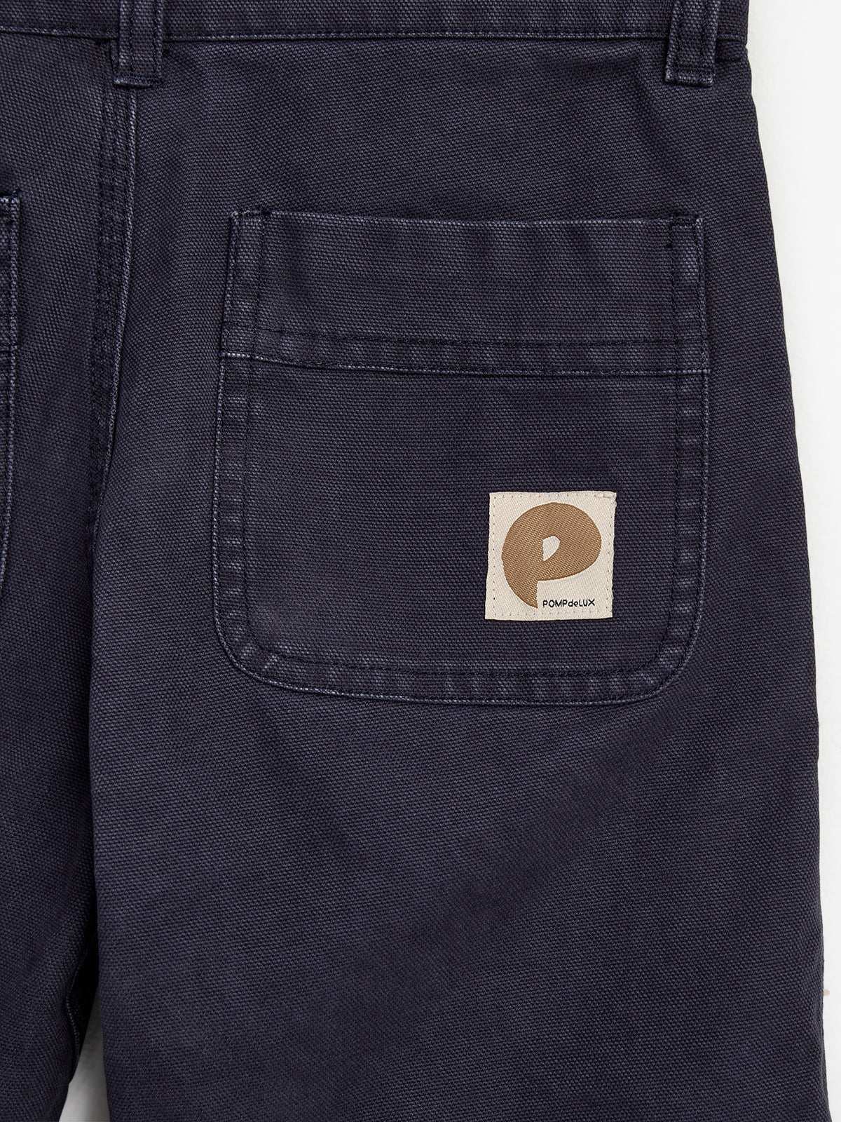 Dark blue pants with a visible back pocket displaying a beige label featuring a stylized "P" and the text "POMPdeLUX." The pants hang on a plain background.
