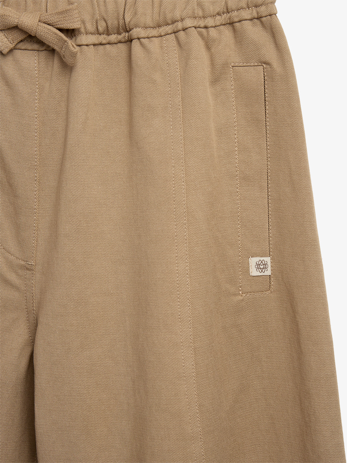 Beige pants with a drawstring waistband and side pocket featuring a small embroidered emblem, set against a plain background.