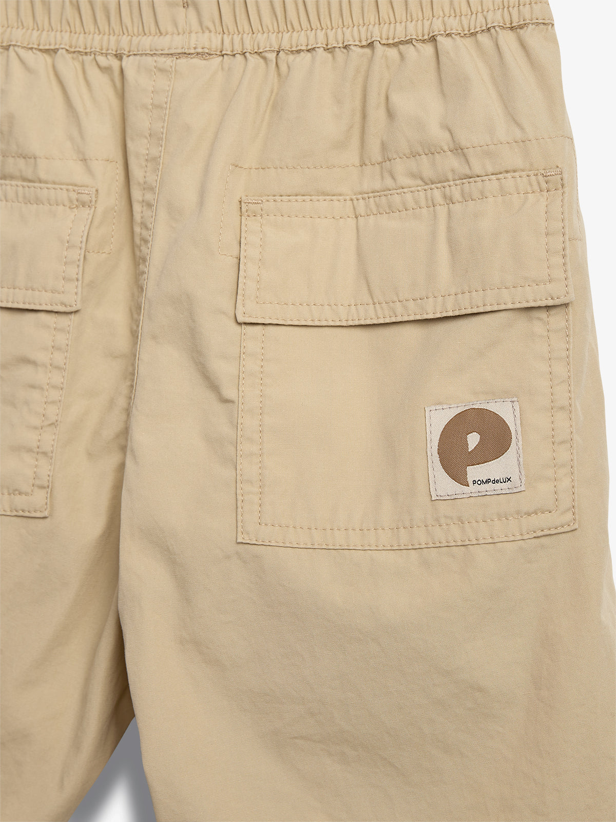 Beige shorts met twee achterzakken, een elastische tailleband en een logo met de tekst "POMPdeLUX" linksonder, tegen een witte achtergrond.
