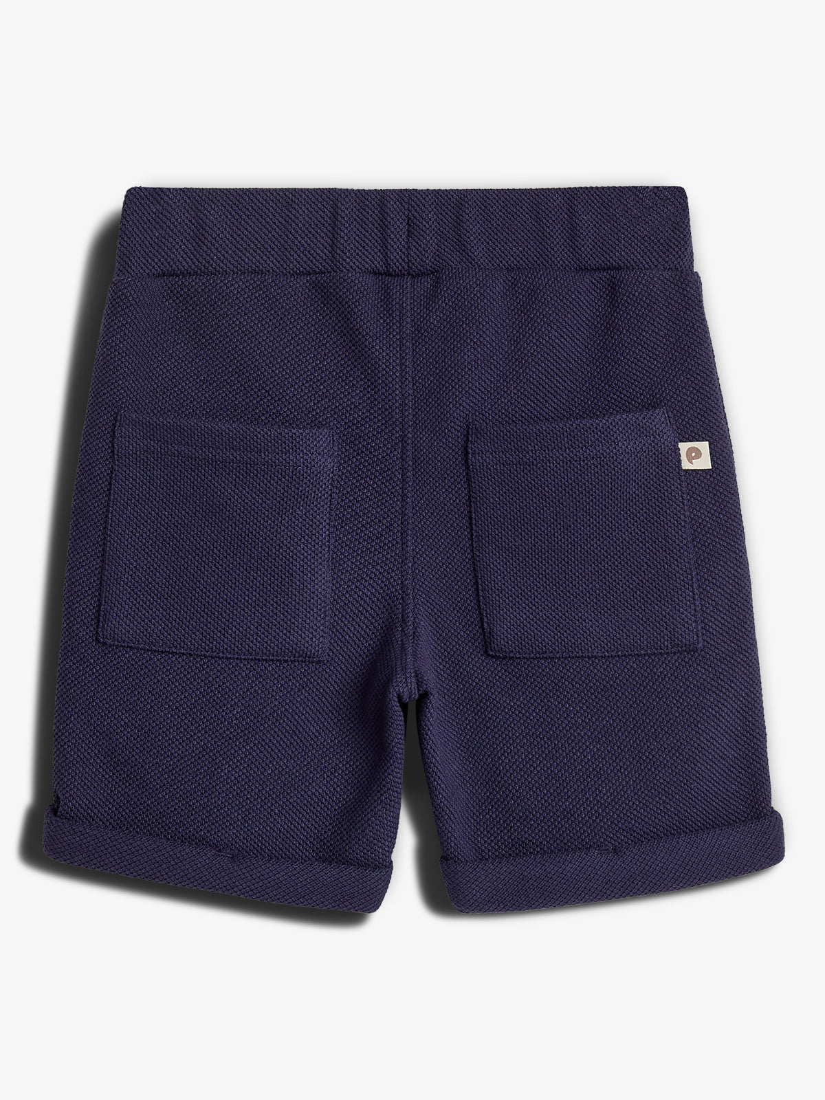 Donkerblauwe korte broek met omgeslagen pijpen en elastische tailleband, twee zakken aan de achterkant. Er is een klein label met een logo aan de rechterzak bevestigd. Achtergrond is wit.