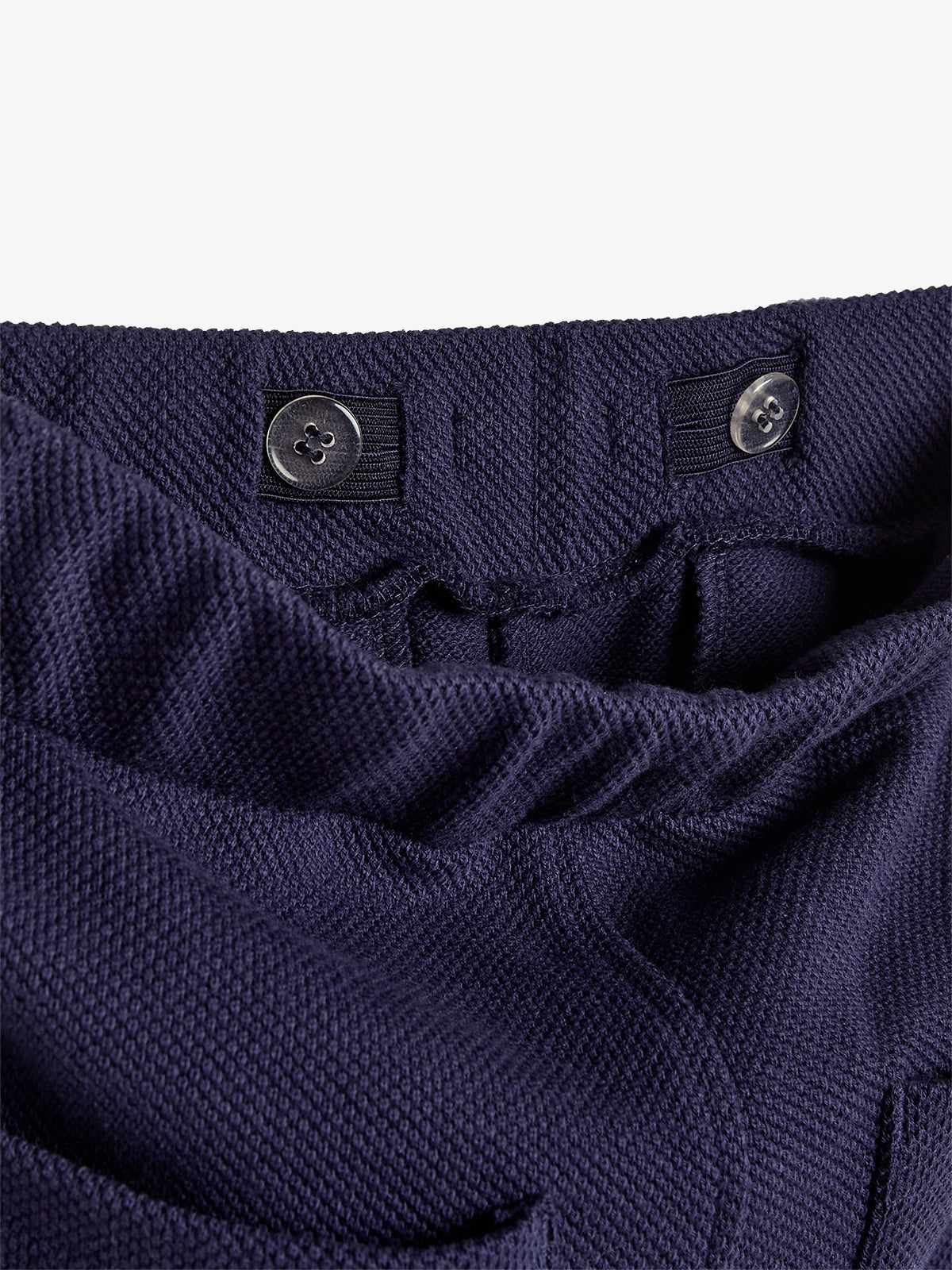 Navy blue textured fabric with an elastic waistband features two adjustable buttons threaded through loops, creating a gathered appearance. The background is a simple off-white surface.