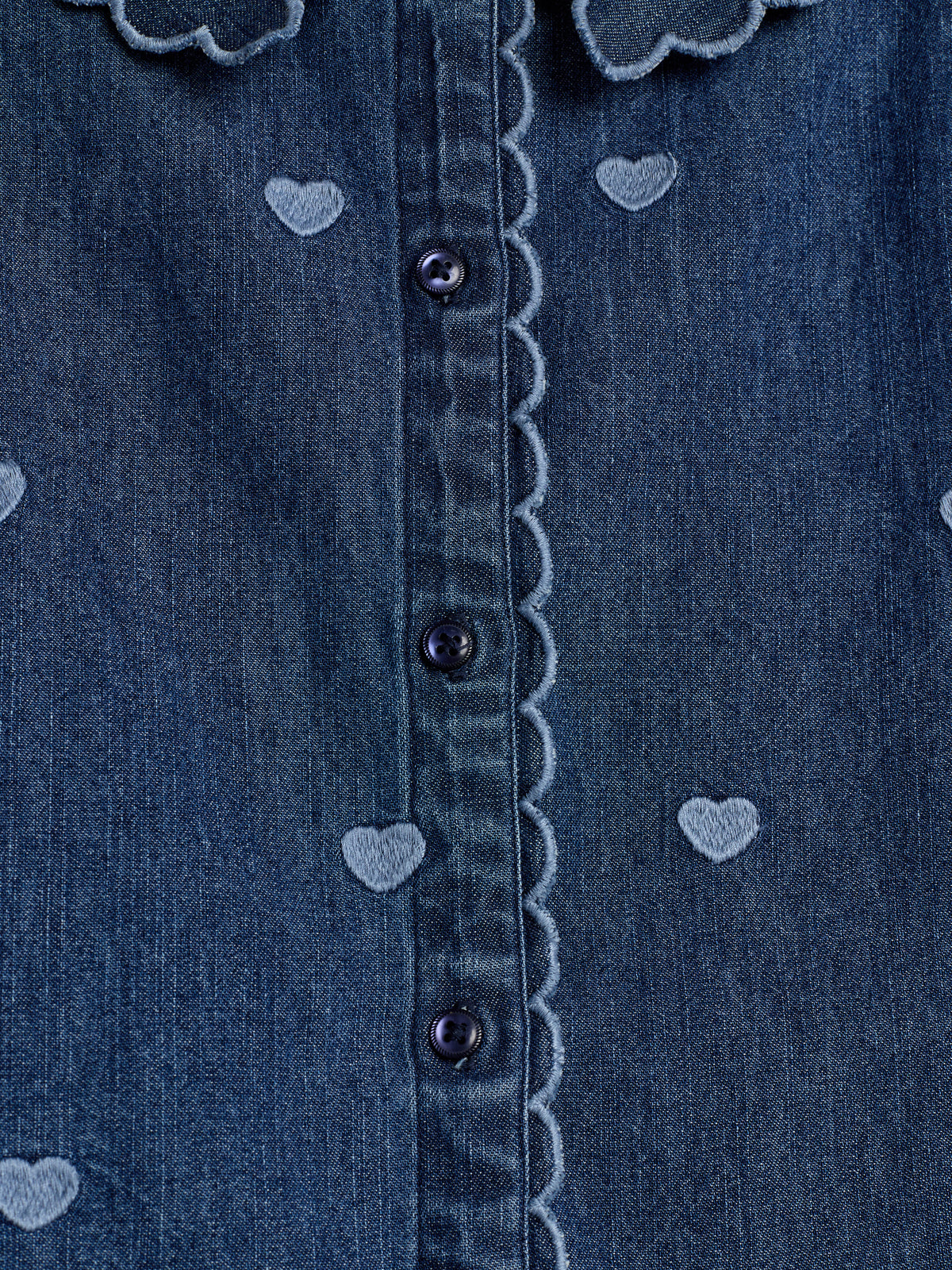 Denim blouse met donkerblauwe knopen accentueert een patroon van geborduurde hartjes. De sierlijke, gegolfde randen voegen detail toe, binnen een eenvoudige achtergrond van effen stof.