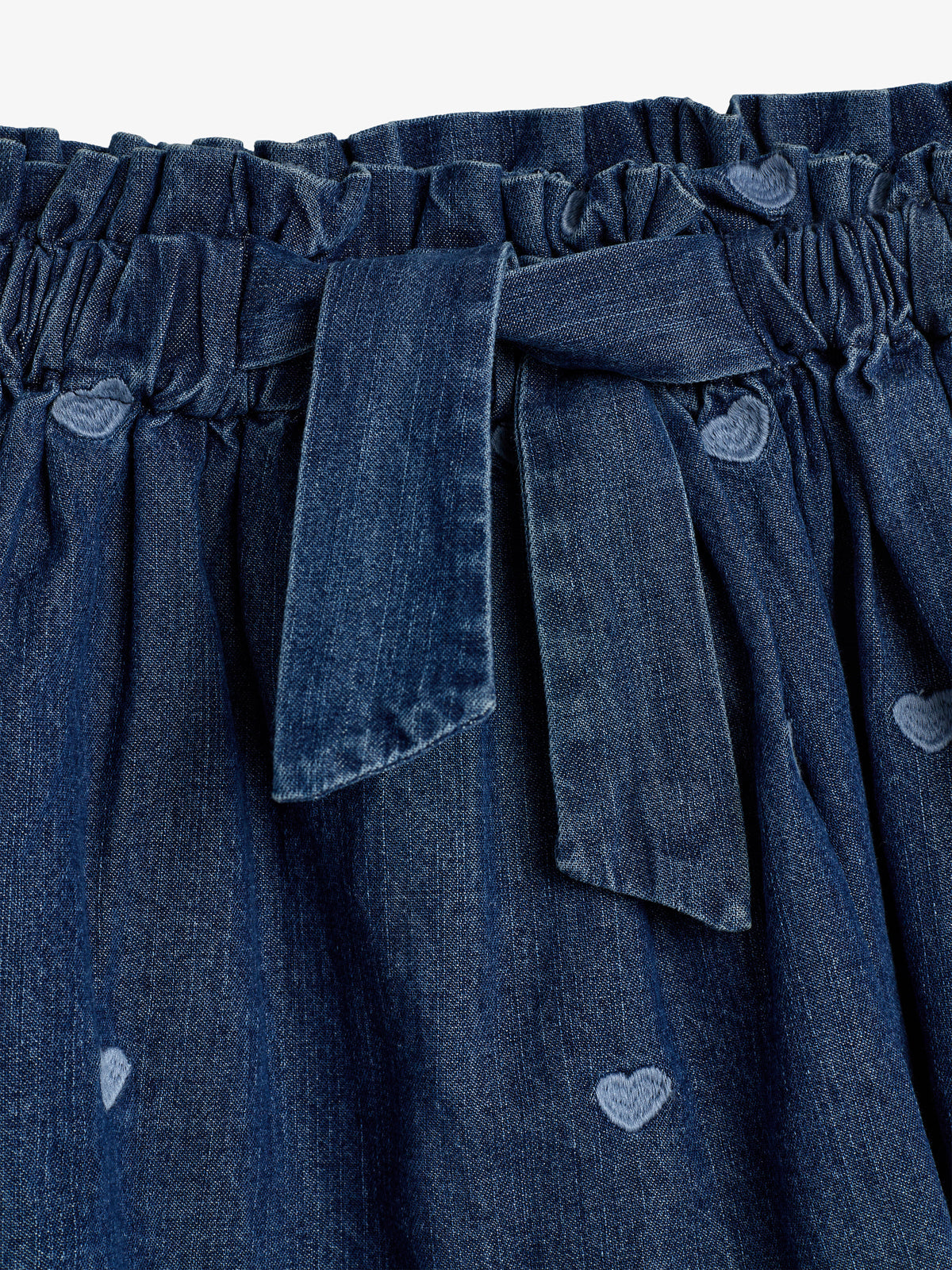 Een blauw denim rok heeft een elastische tailleband met een strik. Hartvormige patronen zijn verspreid over de stof. Het ontwerp is eenvoudig en stijlvol.