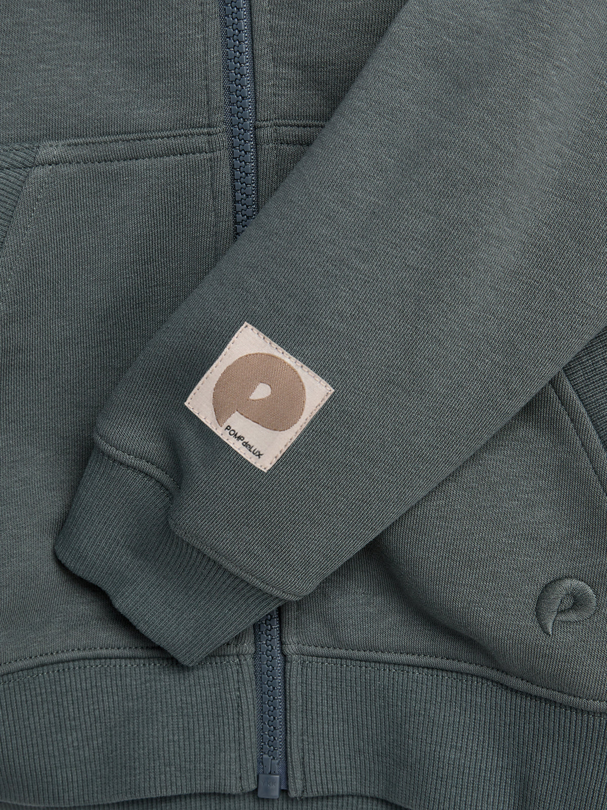 A dark green hoodie sleeve features a small patch with a circular logo and text "POMPELMO." A matching zipper and ribbed fabric design add detail in a casual clothing setting.