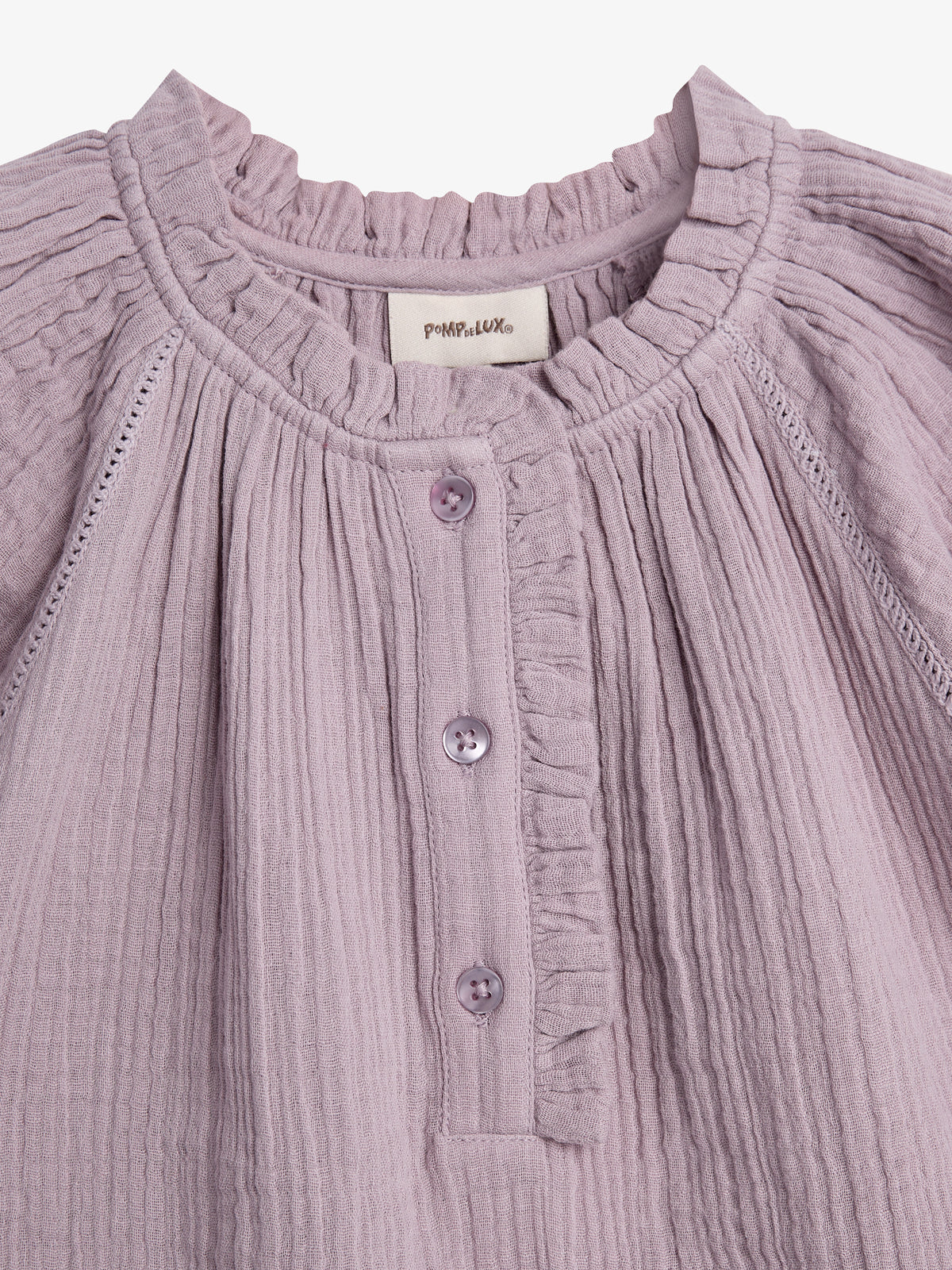 A close-up of a lilac-colored blouse is displayed. The blouse features pleated fabric, a ruffled collar, and a button-up design. A label with "POMPdeLUX" is sewn inside the collar.