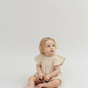 A toddler sits on a light background, wearing a beige dress, holding a toy, and looking upward with an inquisitive expression.