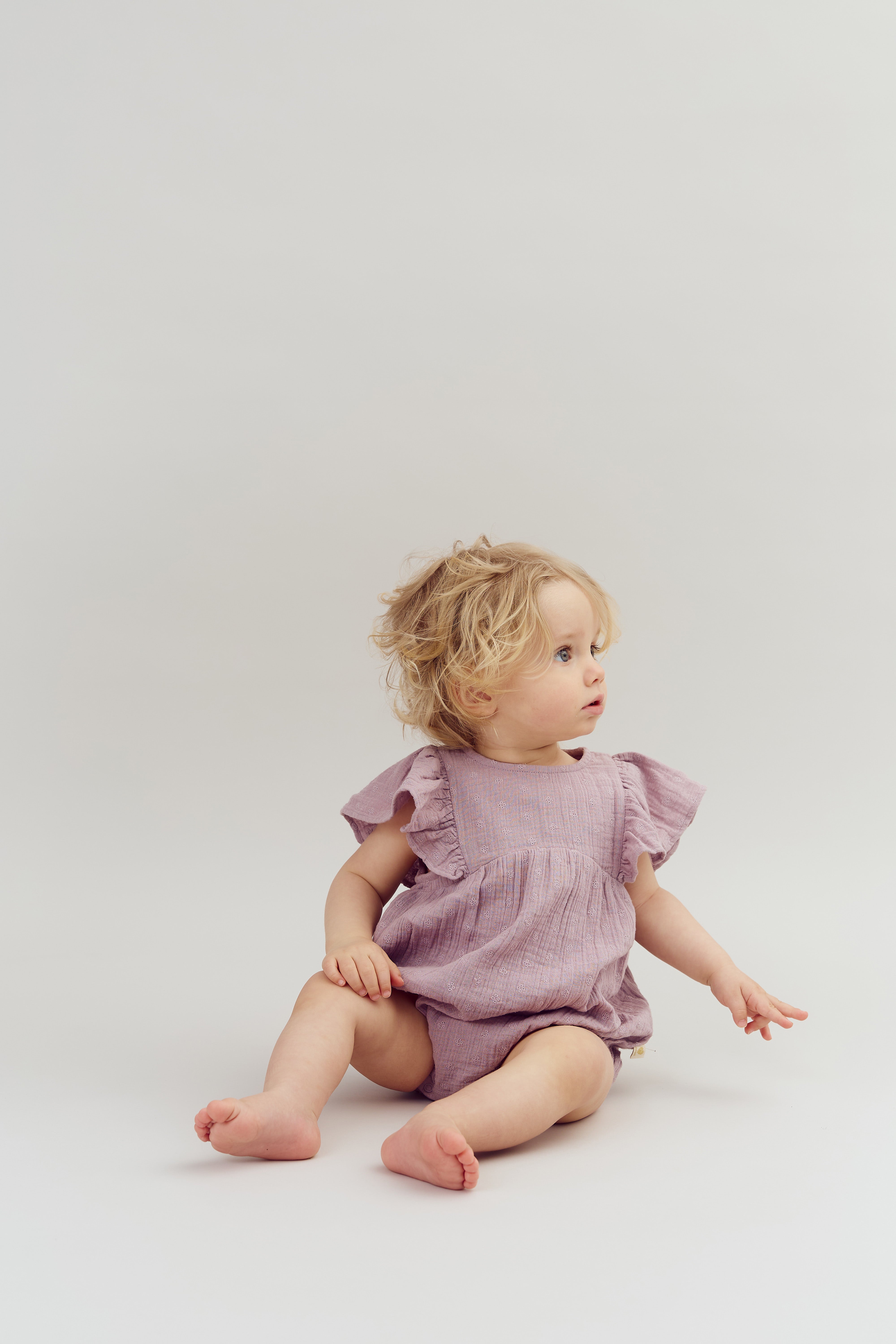 A toddler sits on a plain background, wearing a lavender romper with ruffled sleeves, looking to the side with an attentive expression.