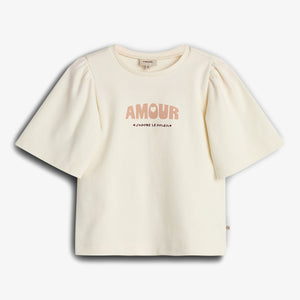 Cream-colored T-shirt featuring puffed sleeves, displays text "AMOUR" and "J’ADORE LE SOLEIL." The shirt is laid flat against a plain white background.