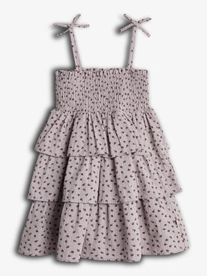 A sleeveless, tiered dress with spaghetti straps and bow detailing. It features a smocked bodice and a floral pattern on a light fabric, set against a plain background.