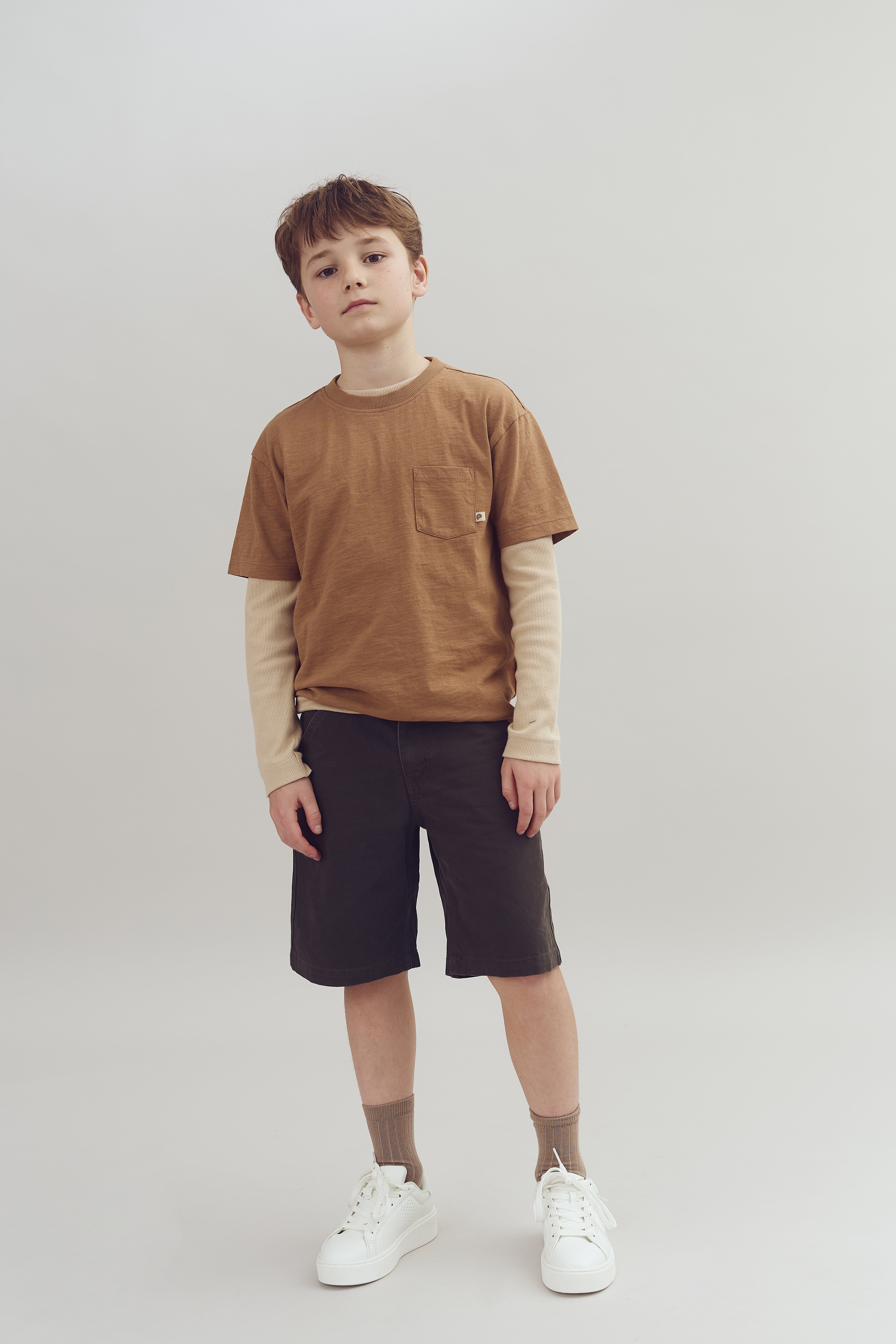 A young boy stands casually, wearing layered beige and brown clothing with white sneakers, in front of a plain gray background.