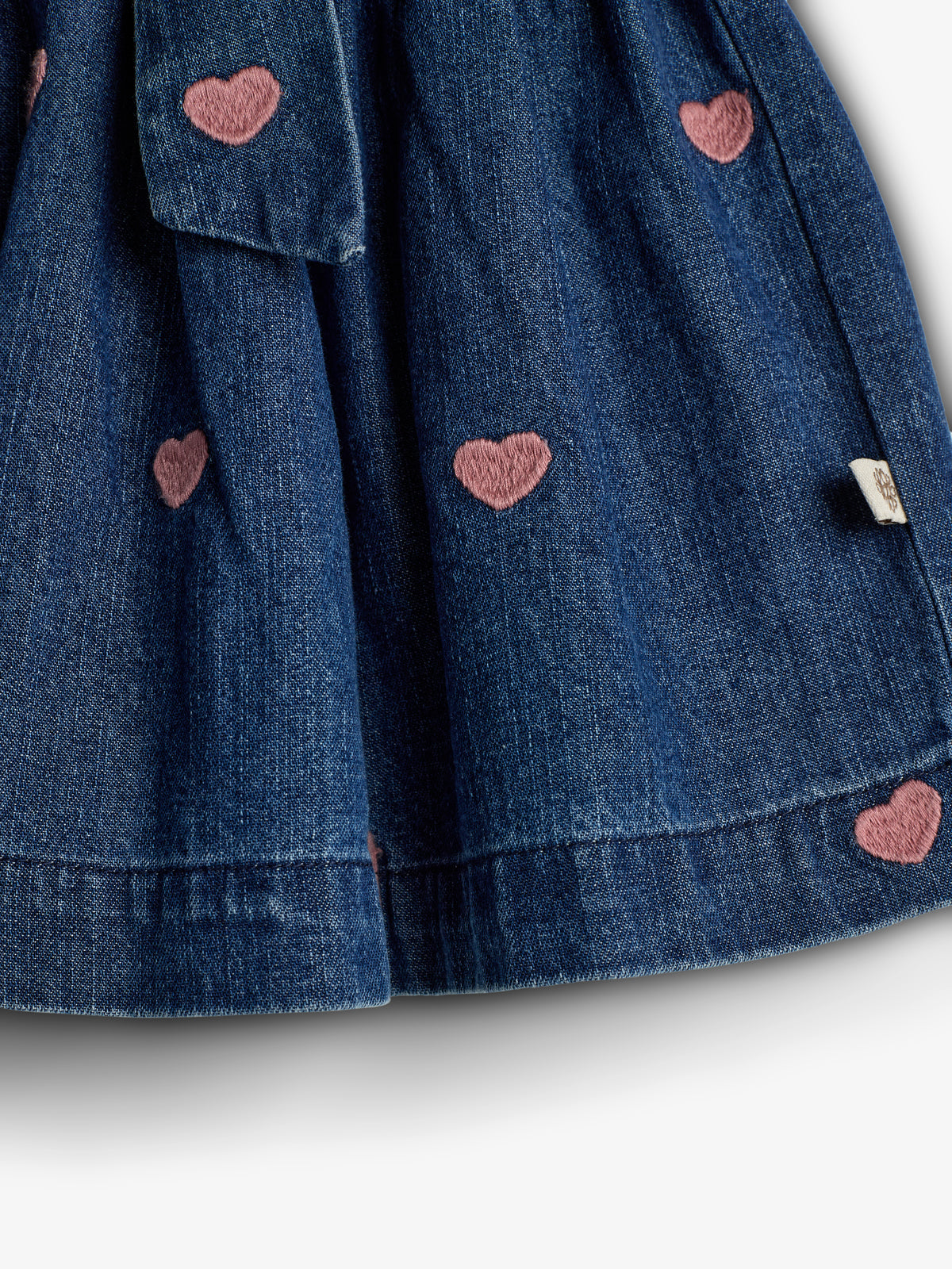 A blue denim fabric with pink heart embroidery scattered throughout. A small tag is attached to the hem, and the fabric appears gathered, suggesting a garment detail.