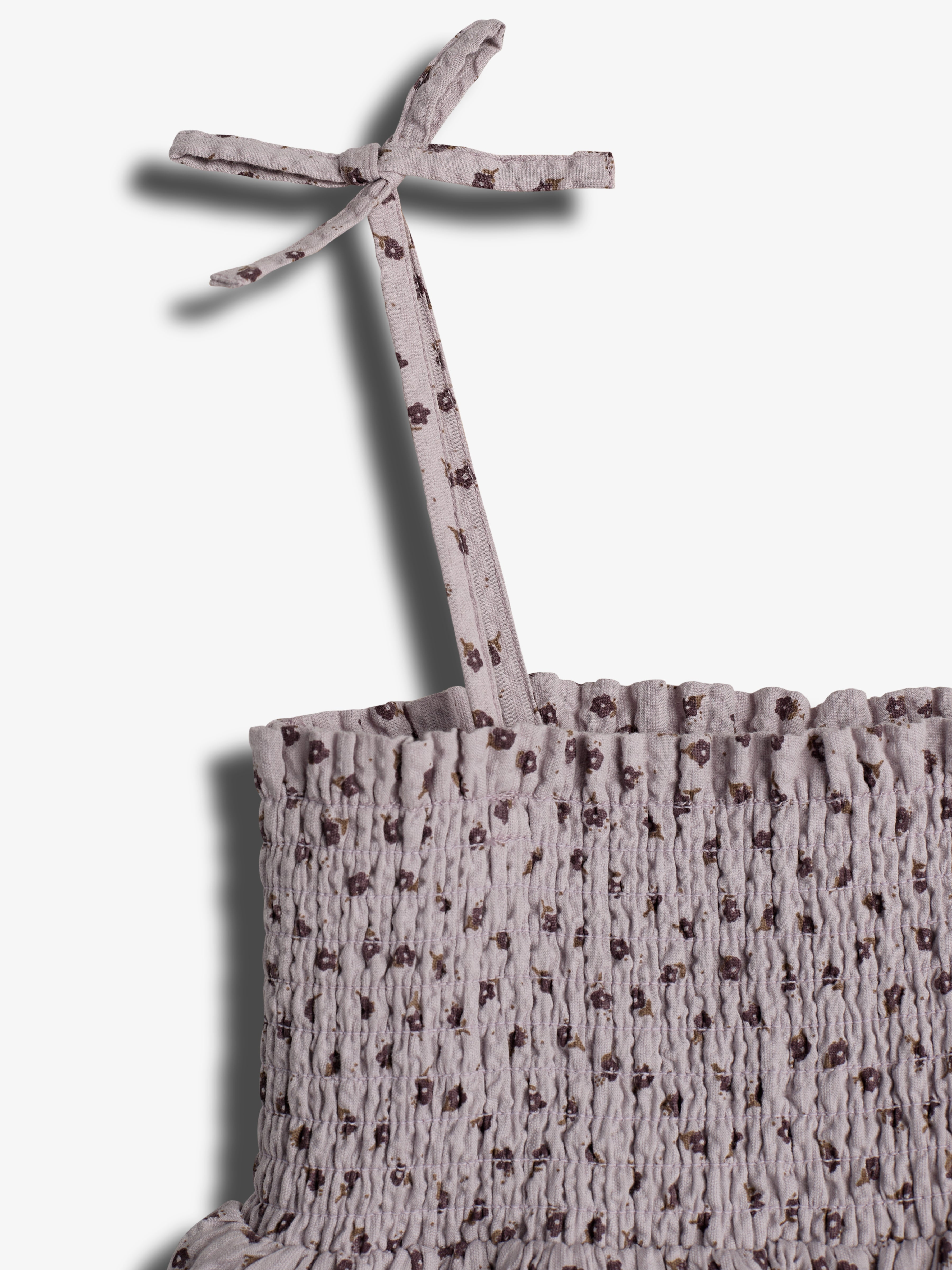 A patterned, pale purple top with a textured, gathered fabric and a tied strap, displayed against a plain white background. The strap features small, dark floral designs.