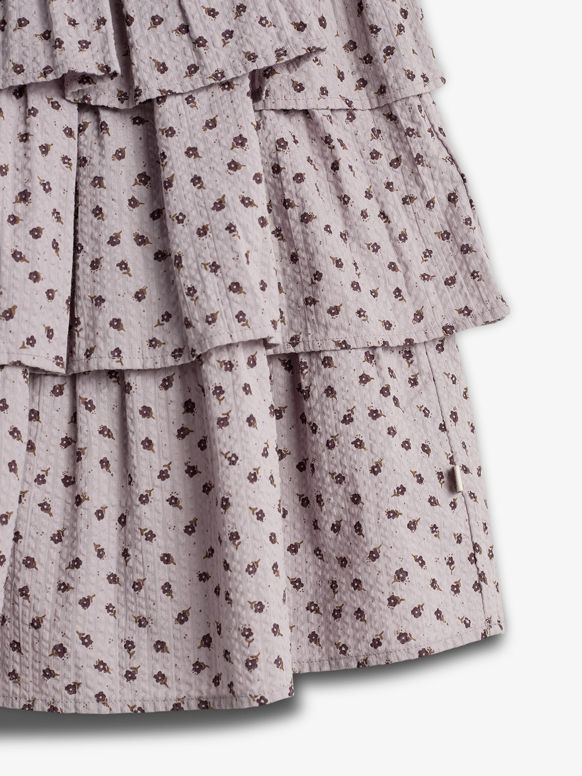 A tiered skirt displays a lilac fabric with a small, dark floral pattern, creating a textured look. It hangs loosely, without visible background or additional objects.