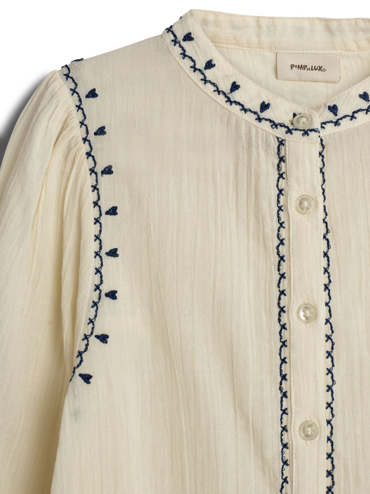 A cream-colored blouse with embroidered navy patterns along the collar and sleeves, featuring buttons down the front. A label inside reads "POMPdeLUX."