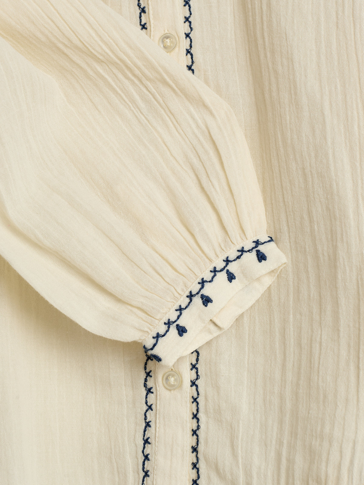 Cream-colored sleeve with blue embroidery and small buttons, featuring a gathered cuff detail. The material has a textured appearance, and the blue stitching adds decorative accents against the fabric.