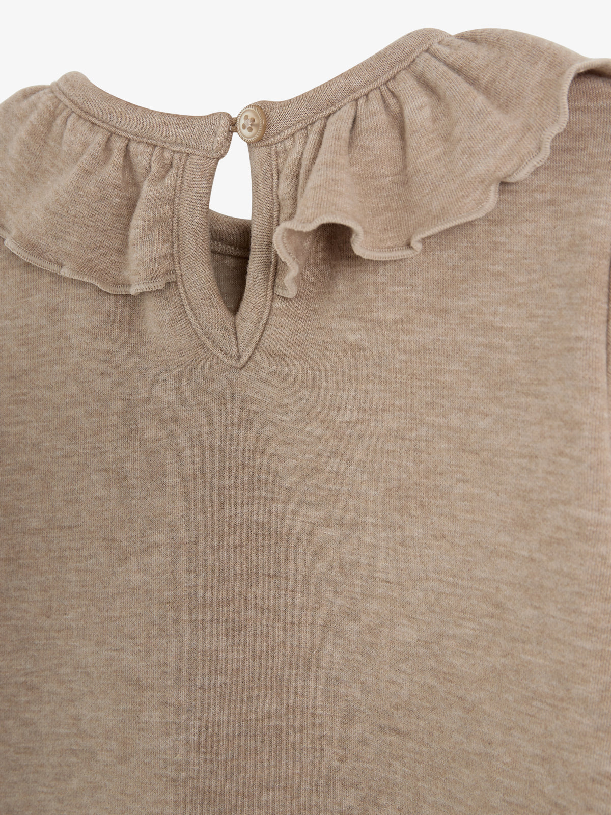 Beige shirt met een rucheskraag en knoopsluiting achter. De stof is zacht en pasteltinten overheersen. De achtergrond is neutraal en egaal van kleur.