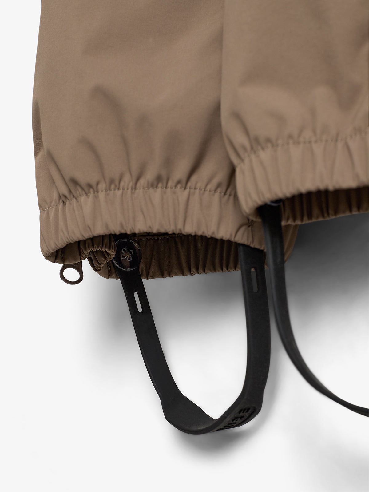 Beige jas met elastische boorden, zichtbaar zijn zwarte verstelbare banden die aan de onderkant hangen. Gelegen tegen een witte achtergrond, de focus ligt op de jasdetails.