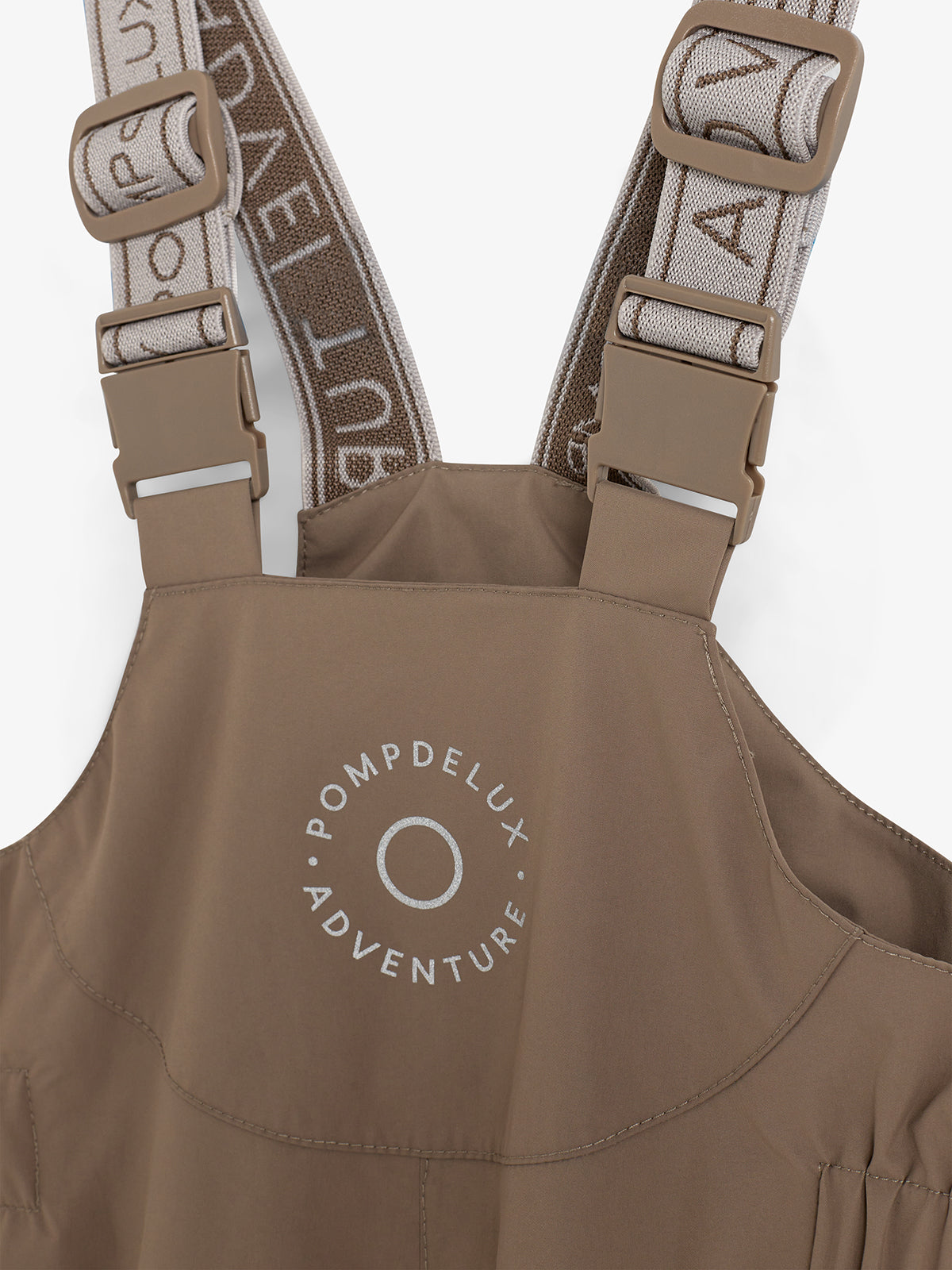 Beige waterdichte broek met bruin gespen, tekst "POMPDELUX ADVENTURE". Twee verstelbare schouderbanden met witte letters. Effen achtergrond.