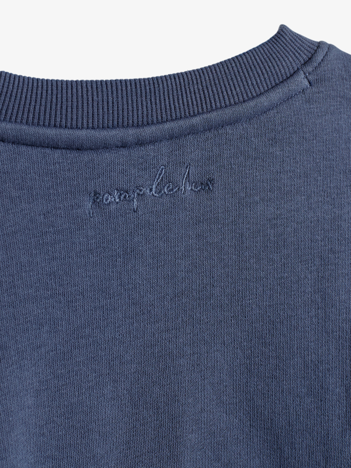 Een blauwe trui hangt rustig; "pompelux" is geborduurd op de kraag. De textuur is zacht en fijngebreid, omgeven door een neutrale achtergrond.
