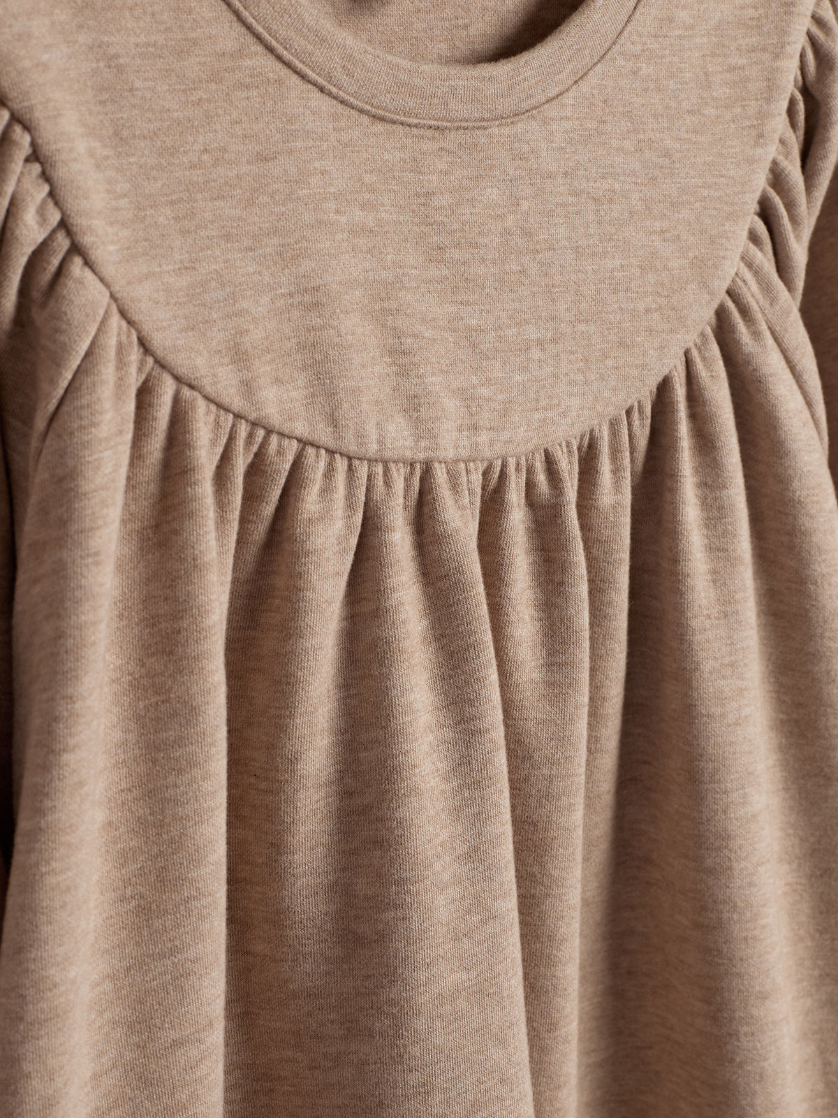 Beige jurk met ronde halslijn en geplooide details, zacht materiaal dat soepel valt, gefotografeerd tegen een neutrale achtergrond.