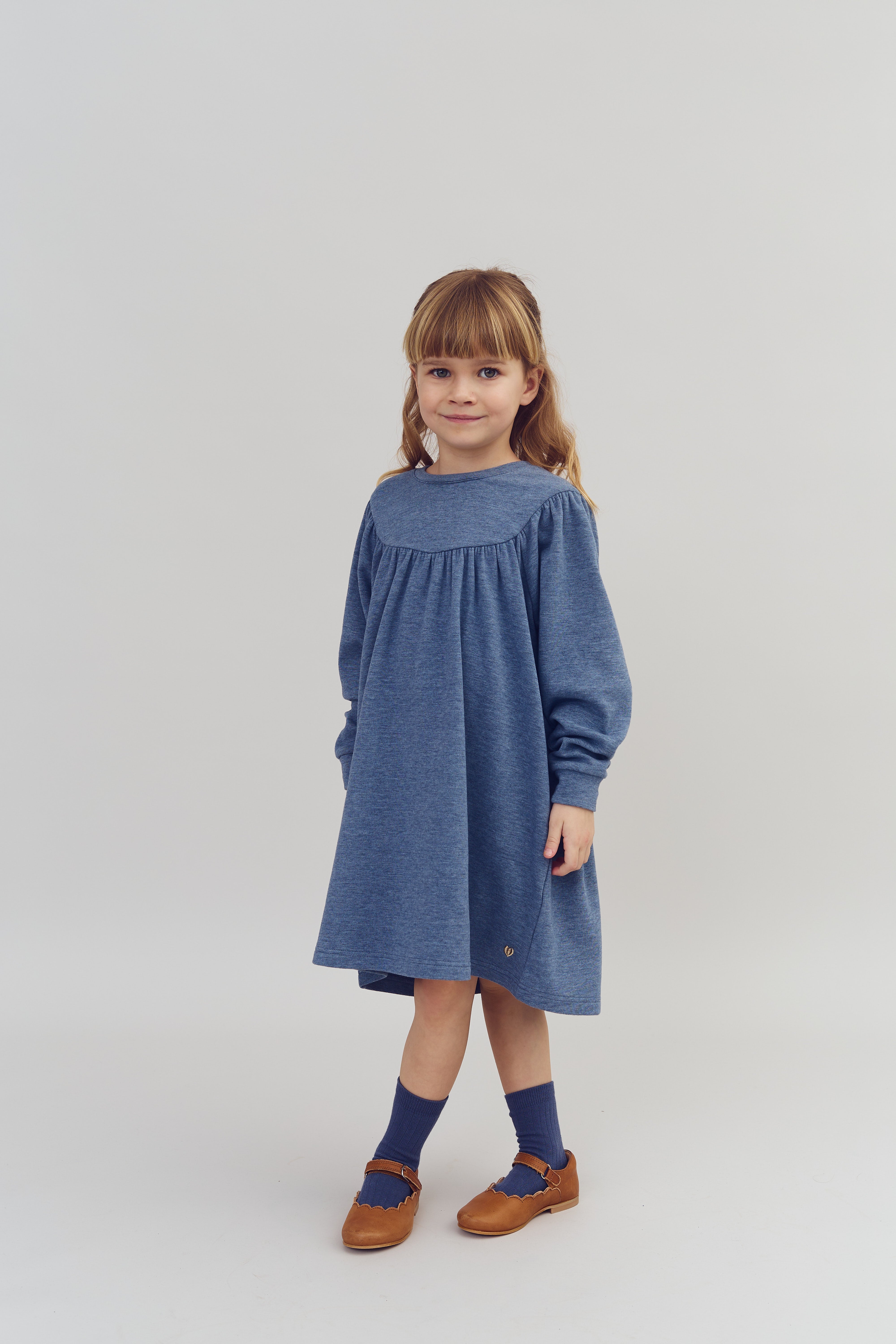 Kind in blauwe jurk staat rechtop, glimlachend in een eenvoudige, lichte studio-omgeving met neutrale achtergrond. Ze draagt bruine schoenen en blauwe sokken.