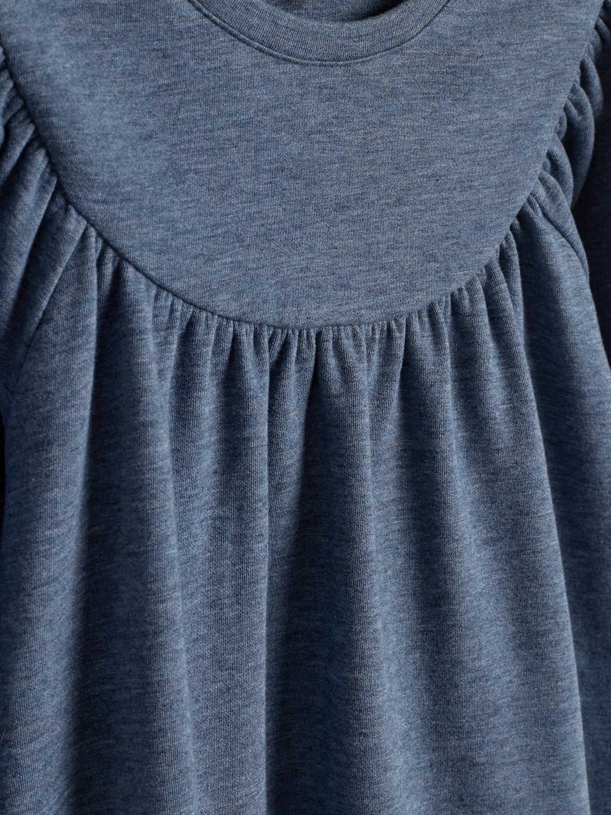 Een blauwgrijs kledingstuk, met plooien onder de ronde halslijn, hangt in neutrale verlichting. De stof lijkt zacht en soepel, geschikt voor casual of comfortabel gebruik.