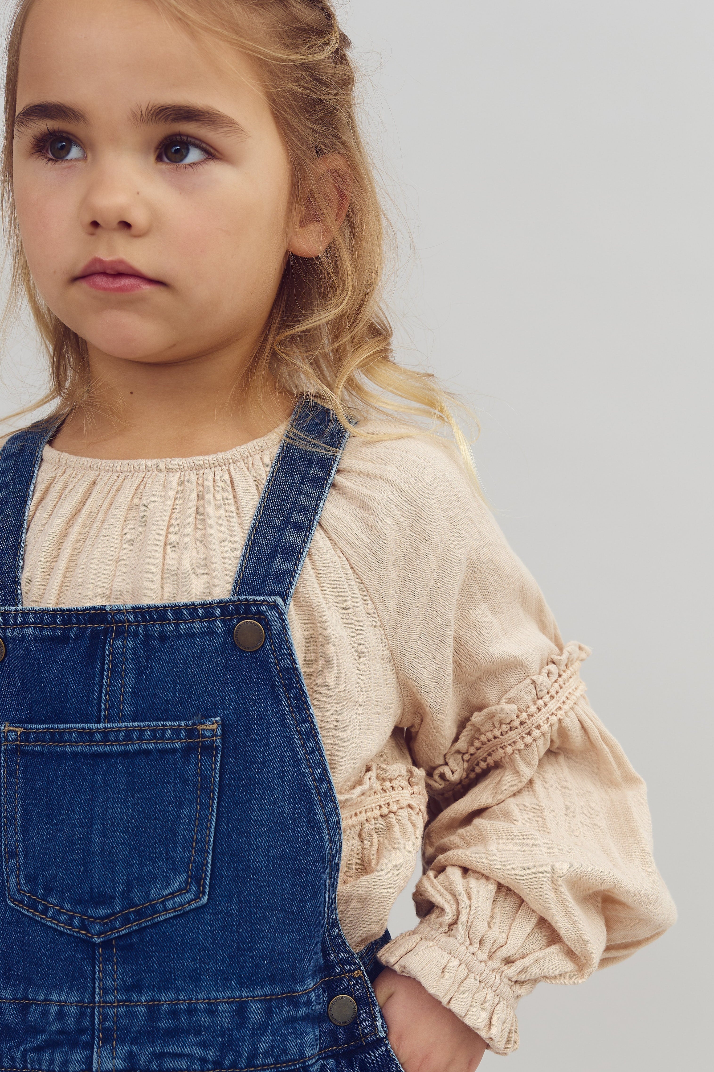 Kind jong meisje draagt een blauwe tuinbroek over een beige blouse en kijkt rustig naar links. Ze heeft lichtbruin haar in een vlecht. De achtergrond is effen grijs.