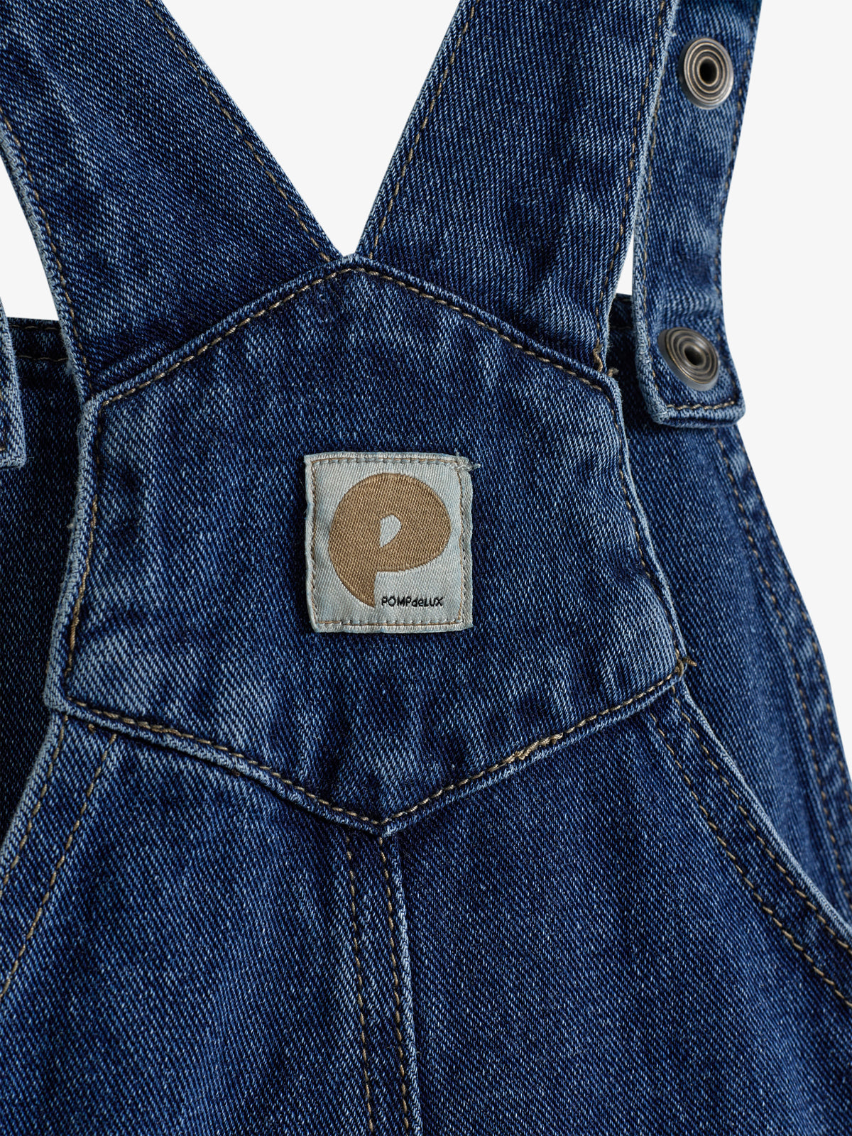 Een donkerblauwe denim tuinbroek met verstelbare bandjes en metalen knopen; een label met logo "POMD'API" is gecentreerd op de borstzak.