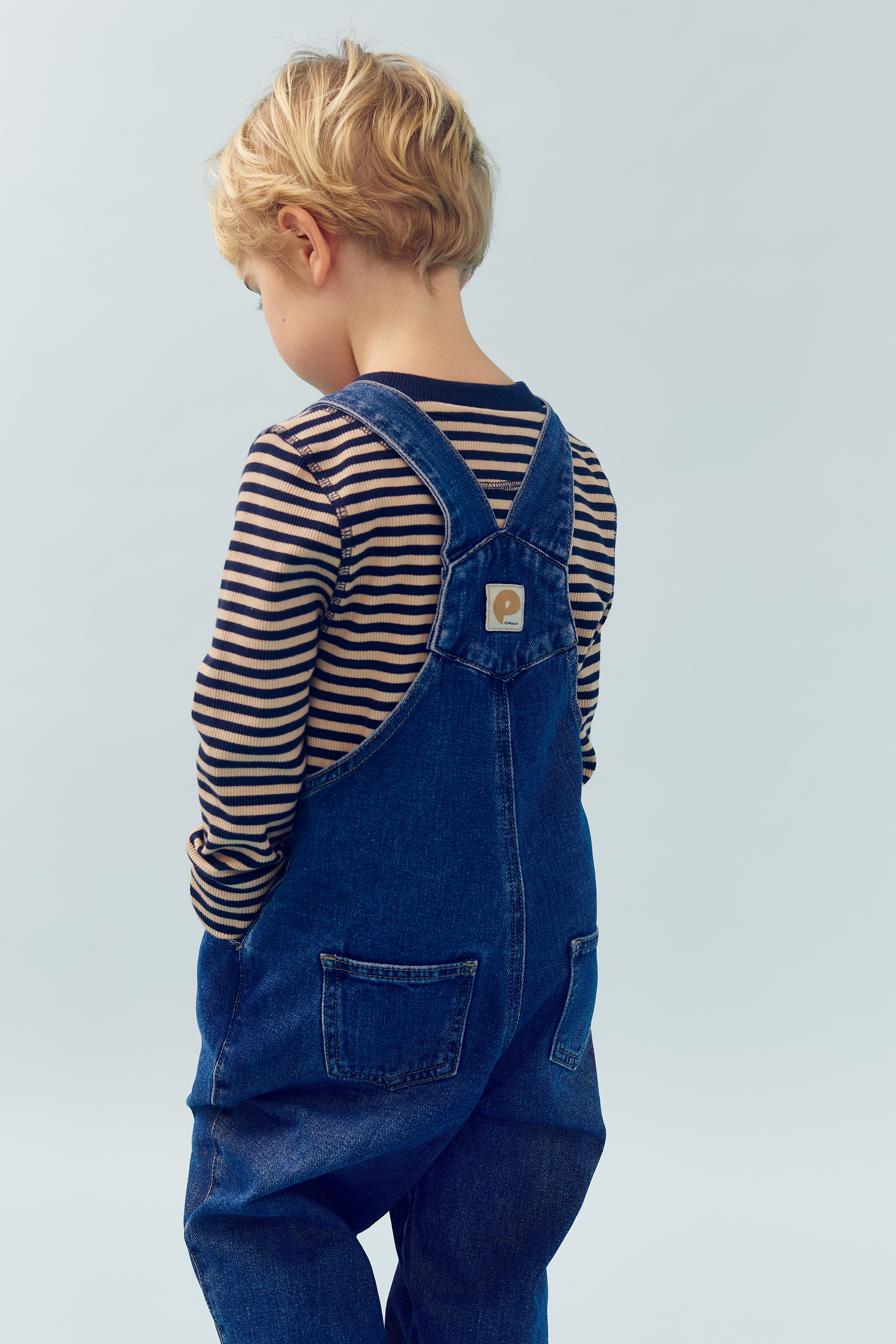 Een kind met blond haar draagt een gestreepte trui en een blauwe tuinbroek. Het kind staat met de rug naar de kijker in een effen omgeving.