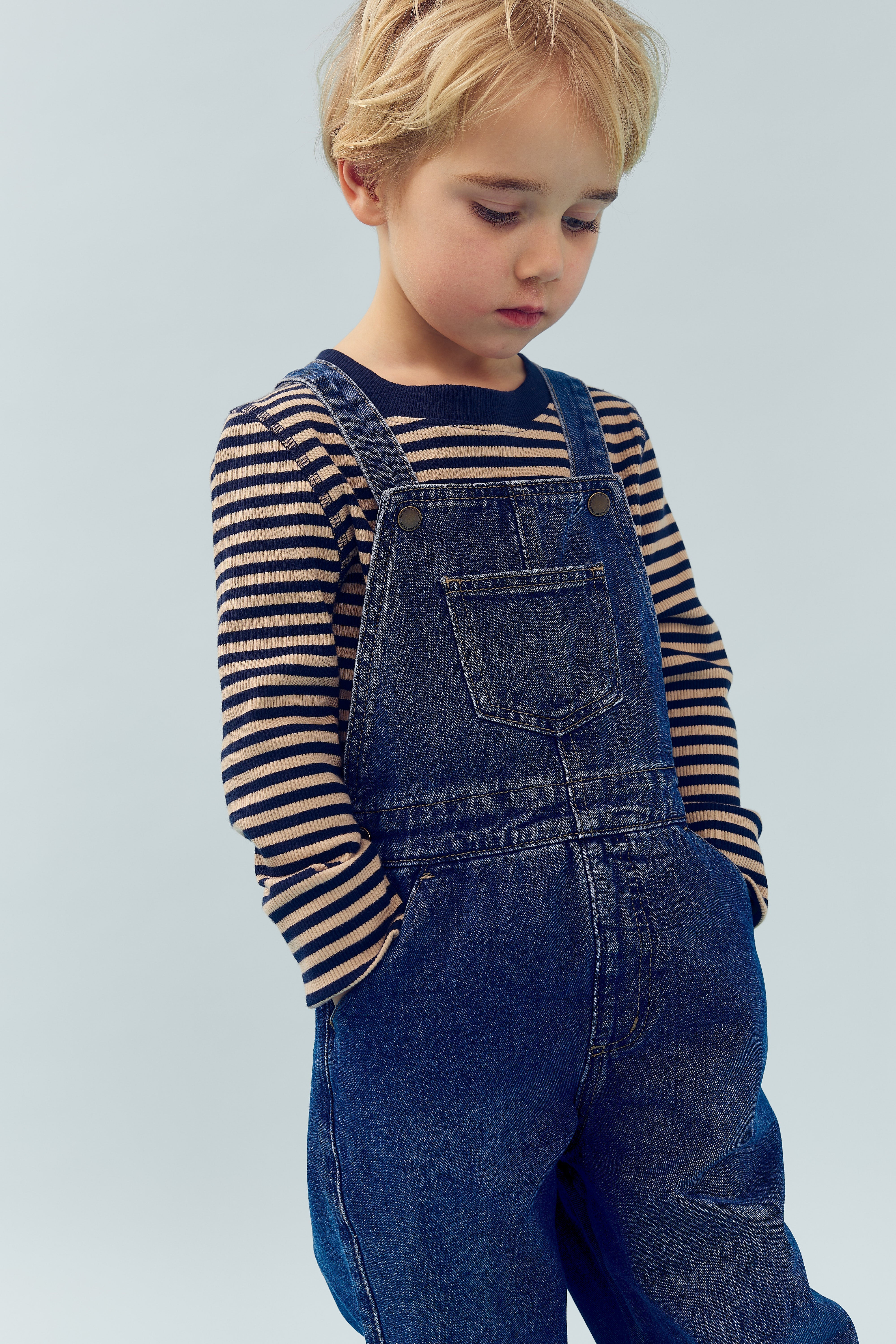 Een kind draagt een blauw gestreepte trui en denim tuinbroek en staat met handen in de zakken tegen een lichte achtergrond, kijkend naar beneden.