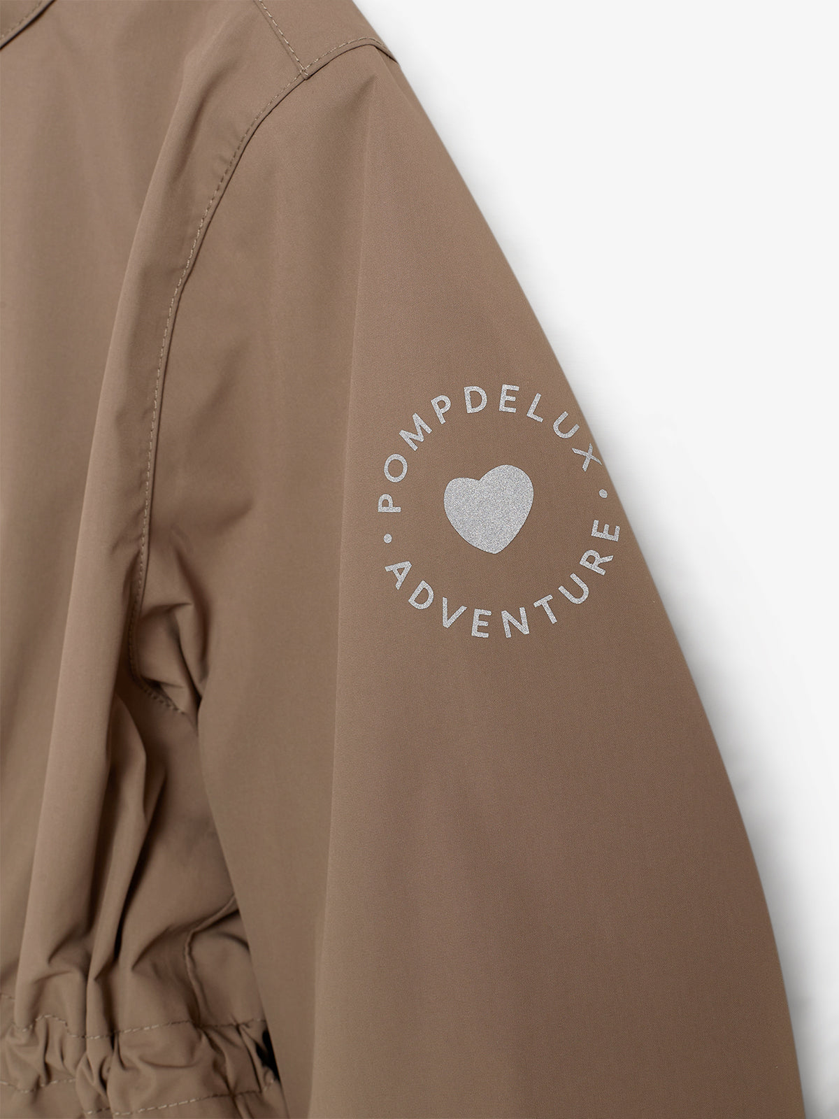 Een bruine jas met opdruk "POMPDELUX ADVENTURE" en een hartje op de mouw, tegen een witte achtergrond. De mouw is onderdeel van de jas en toont plooien en stiksels.