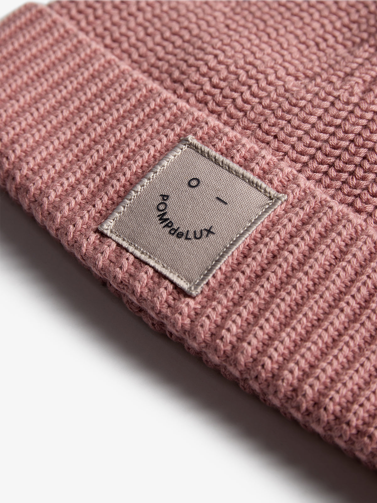 A pink knitted beanie, folded at the brim, features a small gray patch with a simple facial design and text reading "POMPdeLUX" on a white background.