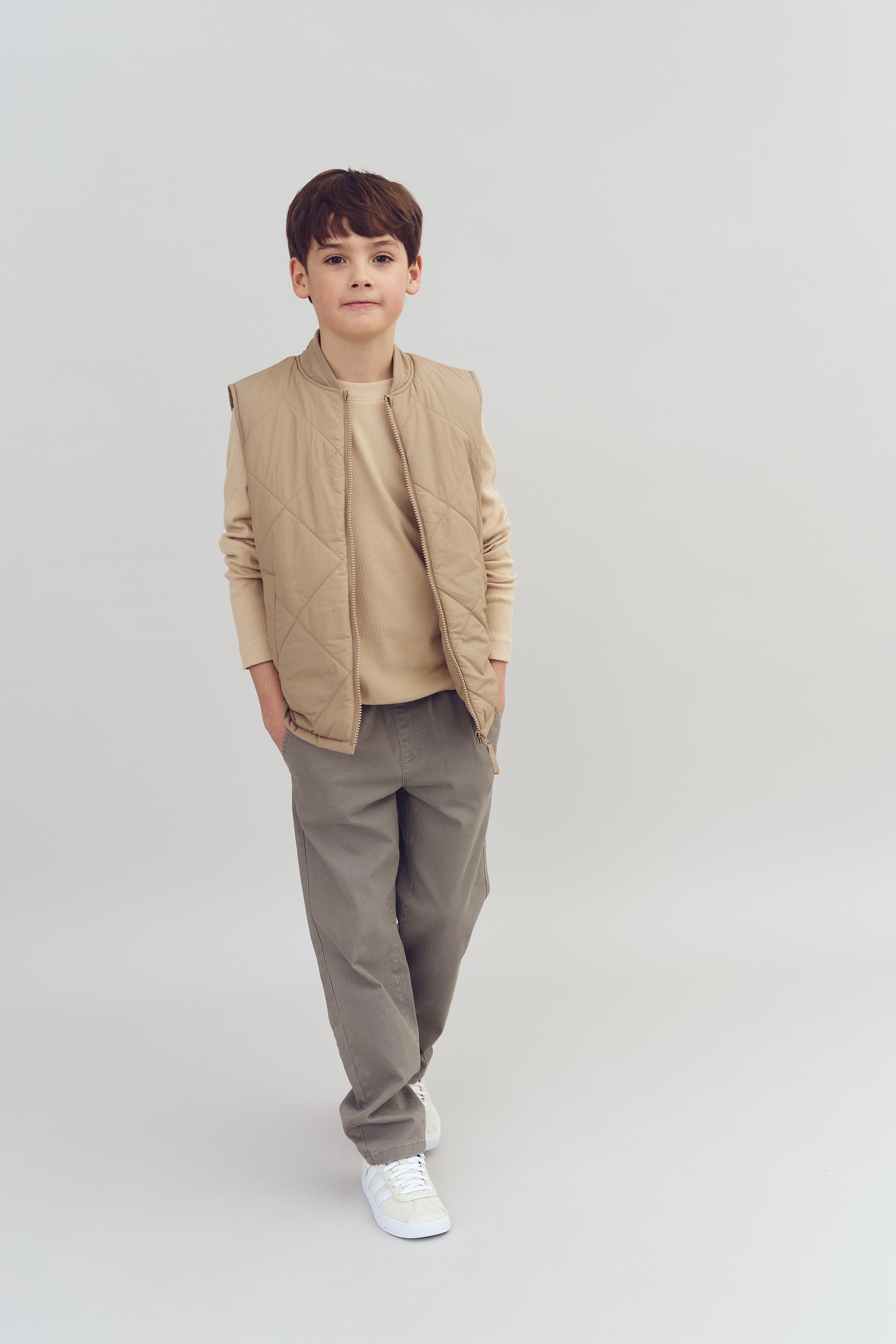Jongen, gekleed in een beige gewatteerd vest en broek, heeft handen in zakken. Hij staat rechtop tegen een neutrale achtergrond.