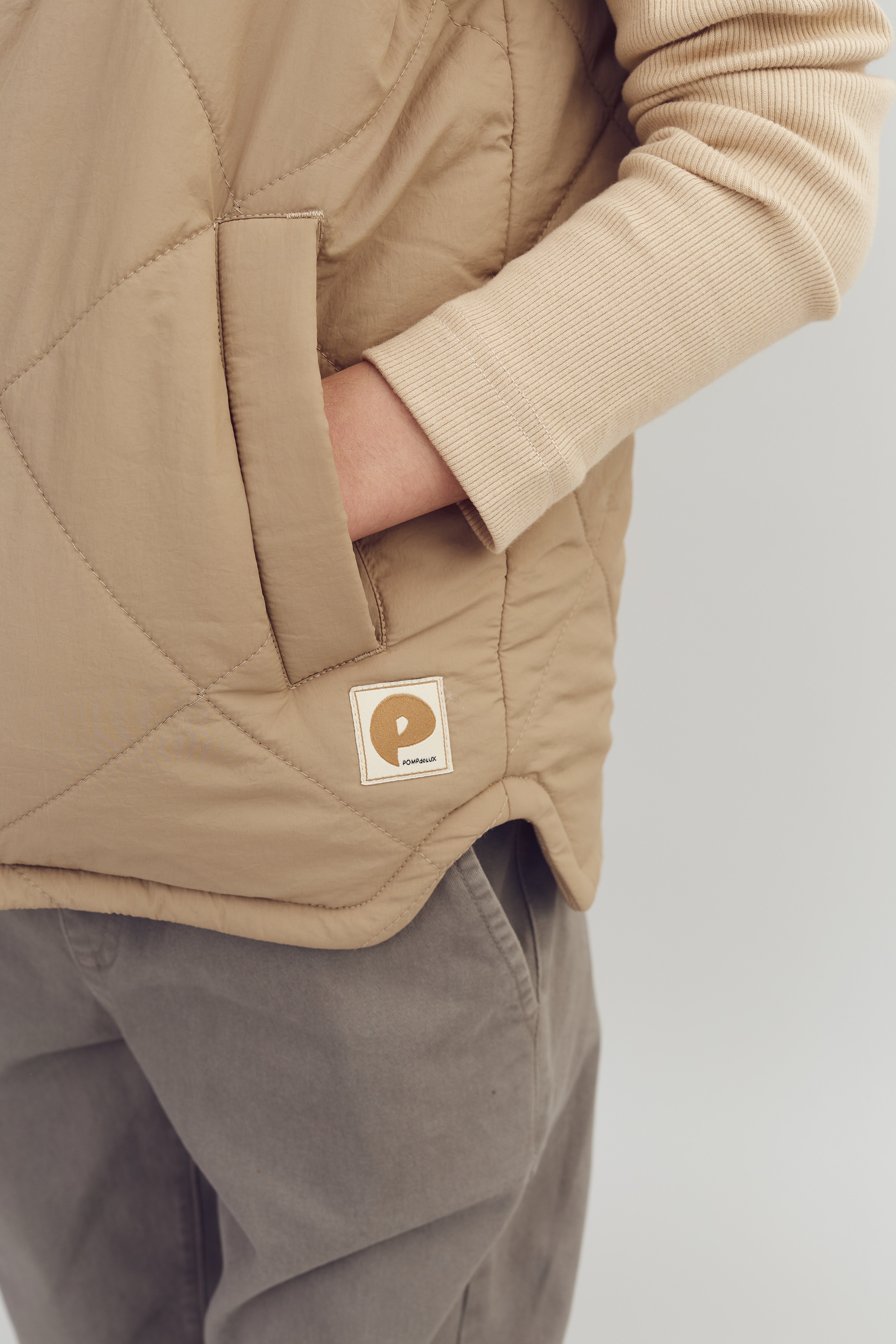 Hand in een beige gewatteerde jaszak, boven een licht geribbelde trui en grijze broek. Label met logo en tekst "POP & SUKI" op de jas. Effen achtergrond.