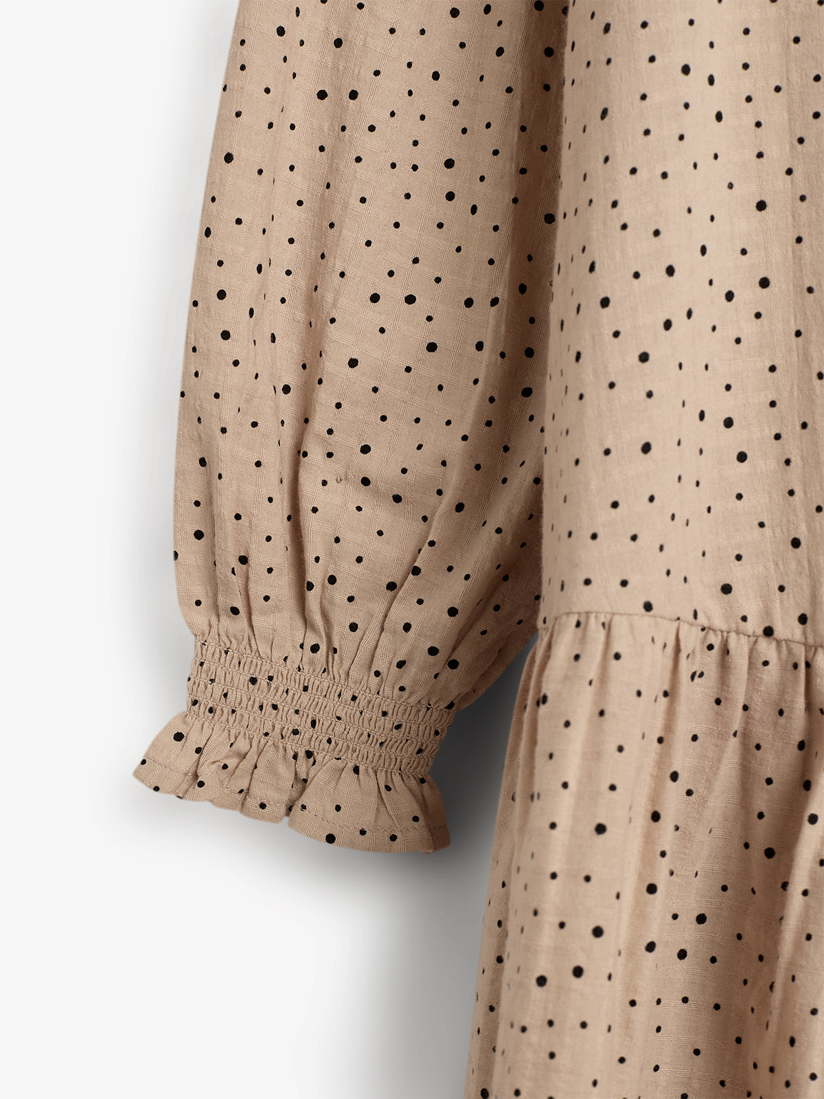 Beige stof met zwarte stippen vormt een kledingstuk dat gevouwen over een achtergrond hangt. De mouw heeft een geplooide manchet, wat een elegante uitstraling geeft.