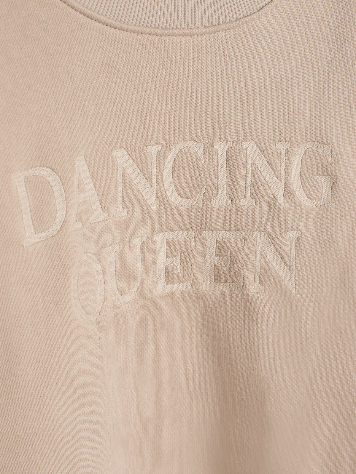 Beige trui met geborduurde tekst "DANCING QUEEN" in een eenvoudige achtergrond, zonder verdere zichtbare elementen.