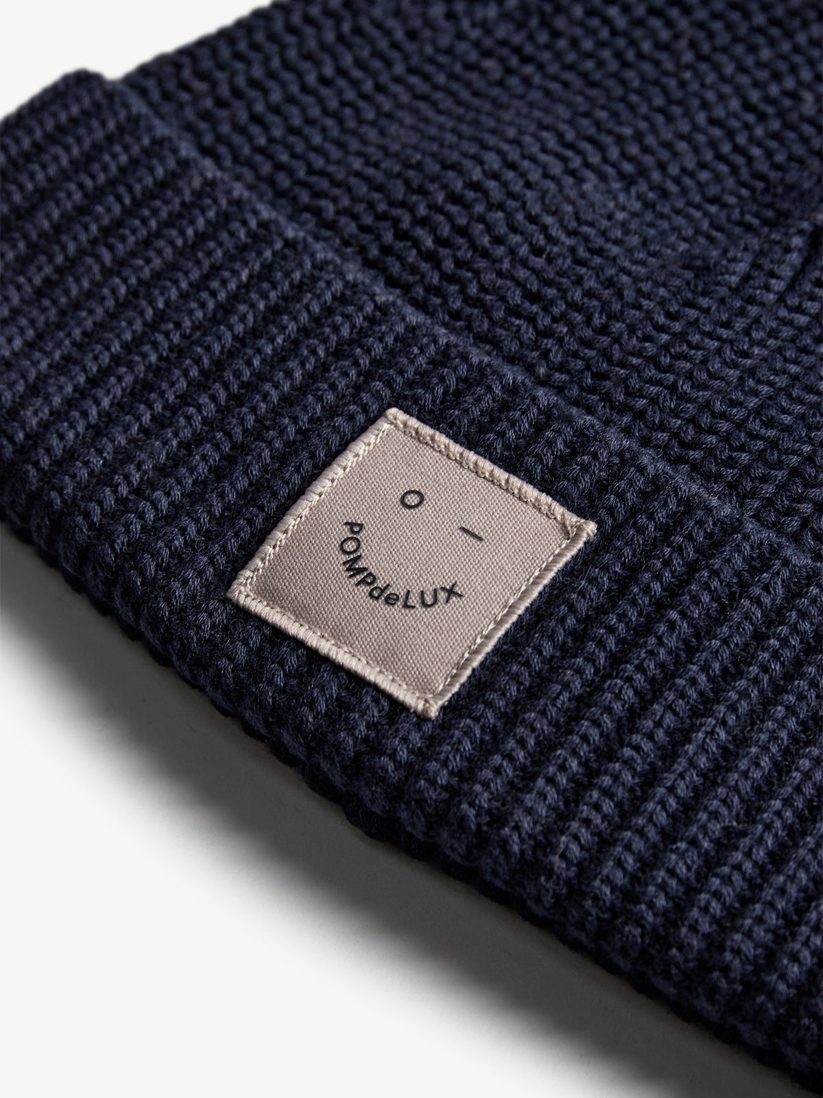A navy knitted beanie features a stitched patch labeled "POmPom de LuX" in a bold font. The beanie lies on a flat, light-colored surface.
