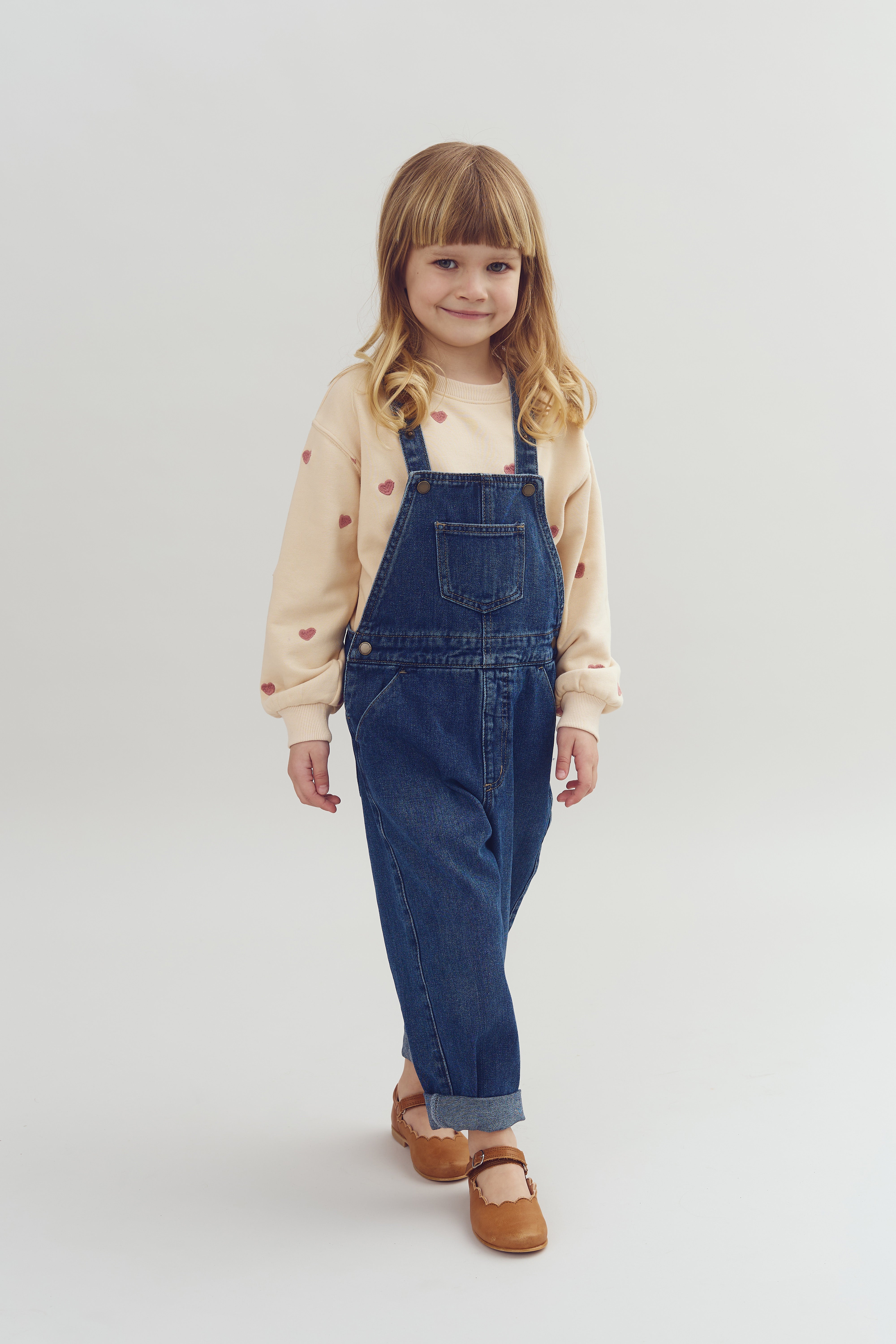 Kind in blauwe tuinbroek en beige trui met hartjes staat glimlachend in een lichte, neutrale ruimte. Ze draagt bruine schoenen en heeft lang, blond haar.