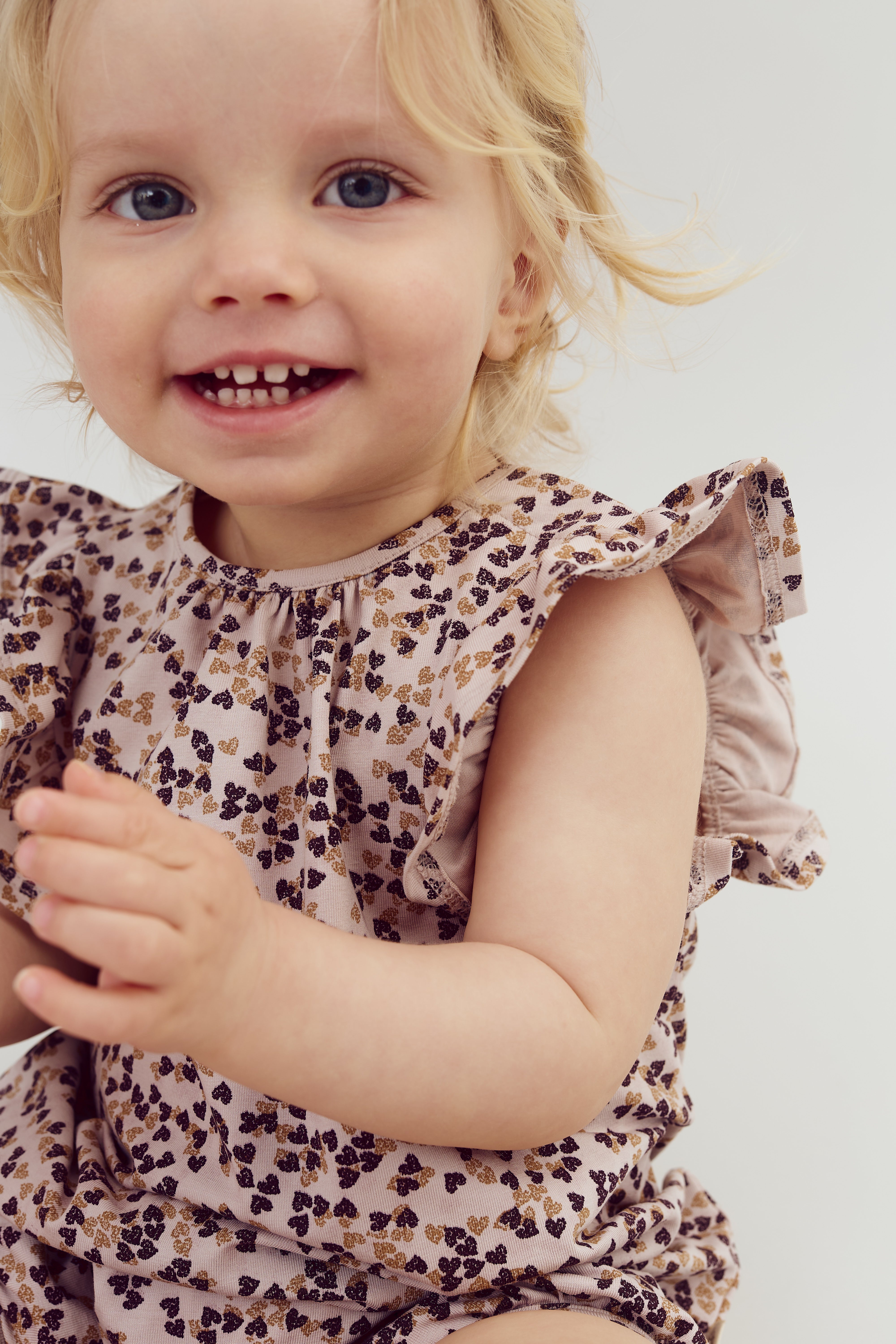 Een peuter met blond haar glimlacht naar de camera. Ze draagt een jurk met een patroon van bruine en gouden blaadjes. De achtergrond is neutraal wit.