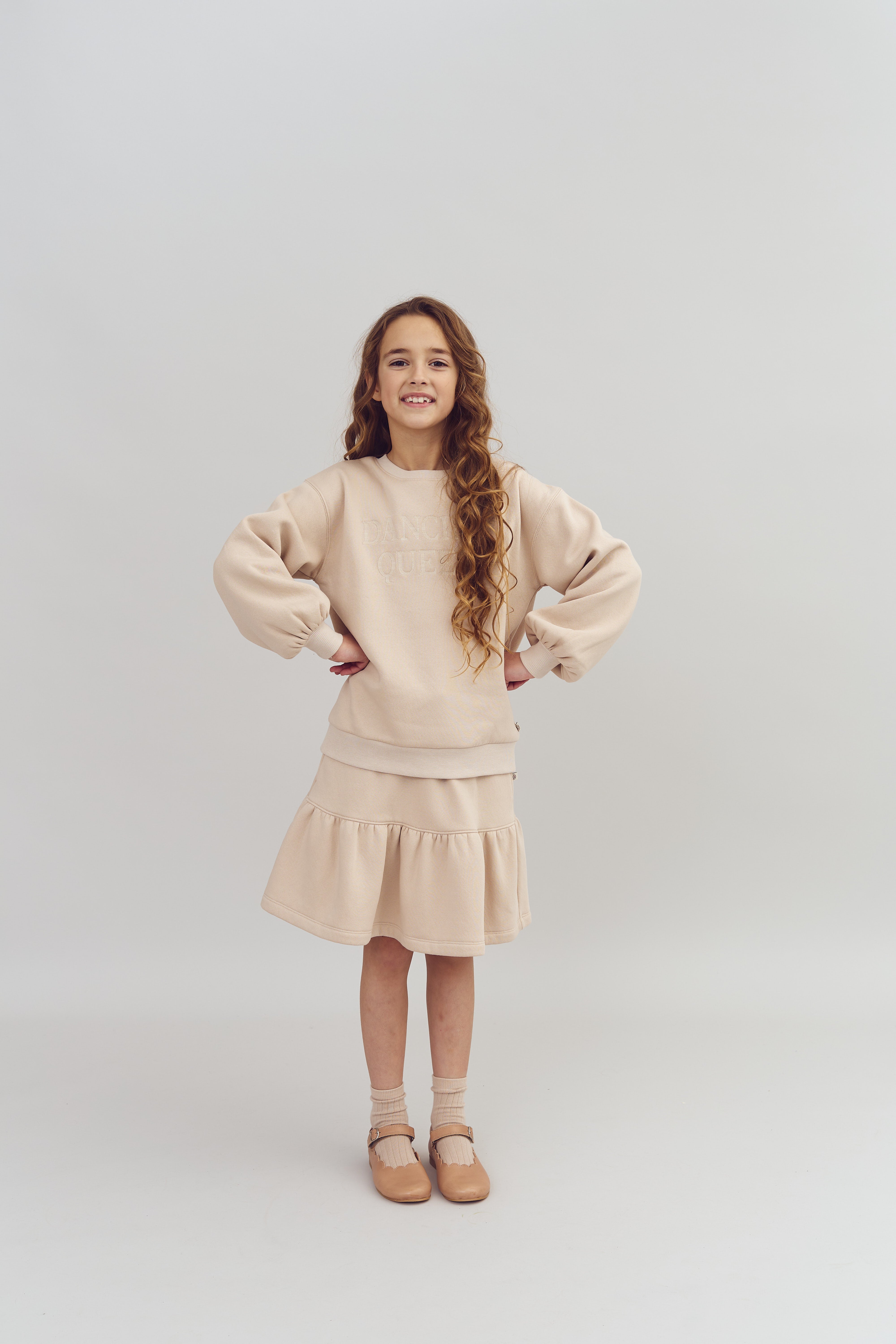 Meisje draagt een beige trui en rok, staat met handen op de heupen, en glimlacht. Achtergrond is neutraal lichtgrijs. Tekst op de trui: "DANCE QUEEN."